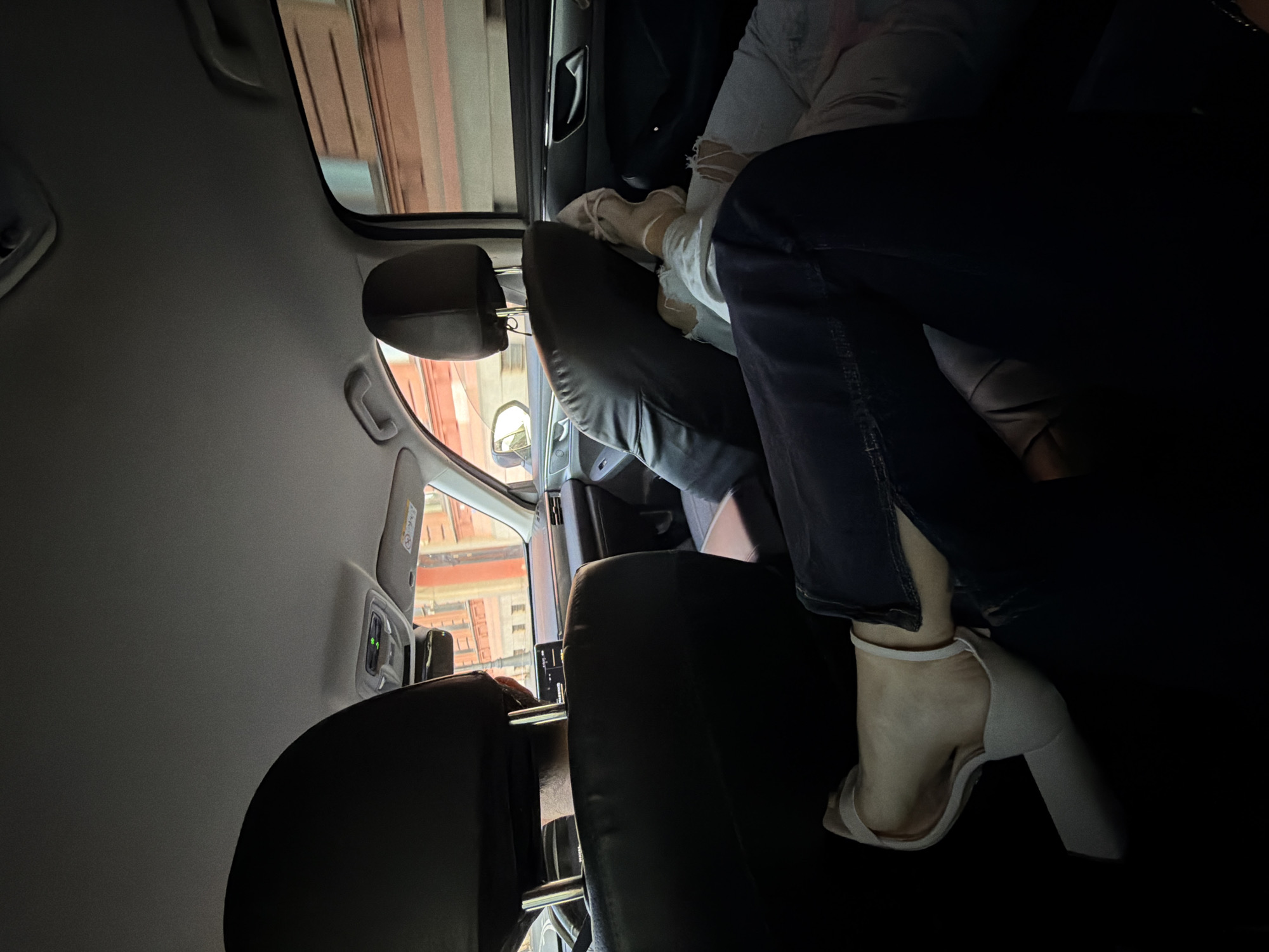 Two individuals are seated in the back of a car, likely a modern sedan, traveling through the city of Saint Petersburg, Russia. The perspective is from the rear passenger seat, looking forward and to the right.
The individual in the foreground, positioned on the left side of the frame, is wearing dark blue denim jeans with a side slit at the hem, revealing a white strap sandal with a block heel. Their legs are casually extended.
The second individual, situated to the right, is wearing light-colored ripped jeans and light-colored shoes. Their legs are also extended, with one foot propped up on a black seatback.
The interior of the car is visible, including the black leather headrests of the front seats, the dashboard with a digital display, and the car's ceiling console with illuminated green buttons. Outside the car, through the windows, the blurred architecture of Saint Petersburg is apparent, featuring buildings with reddish-brown brickwork. The scene suggests a relaxed atmosphere during a car journey.
