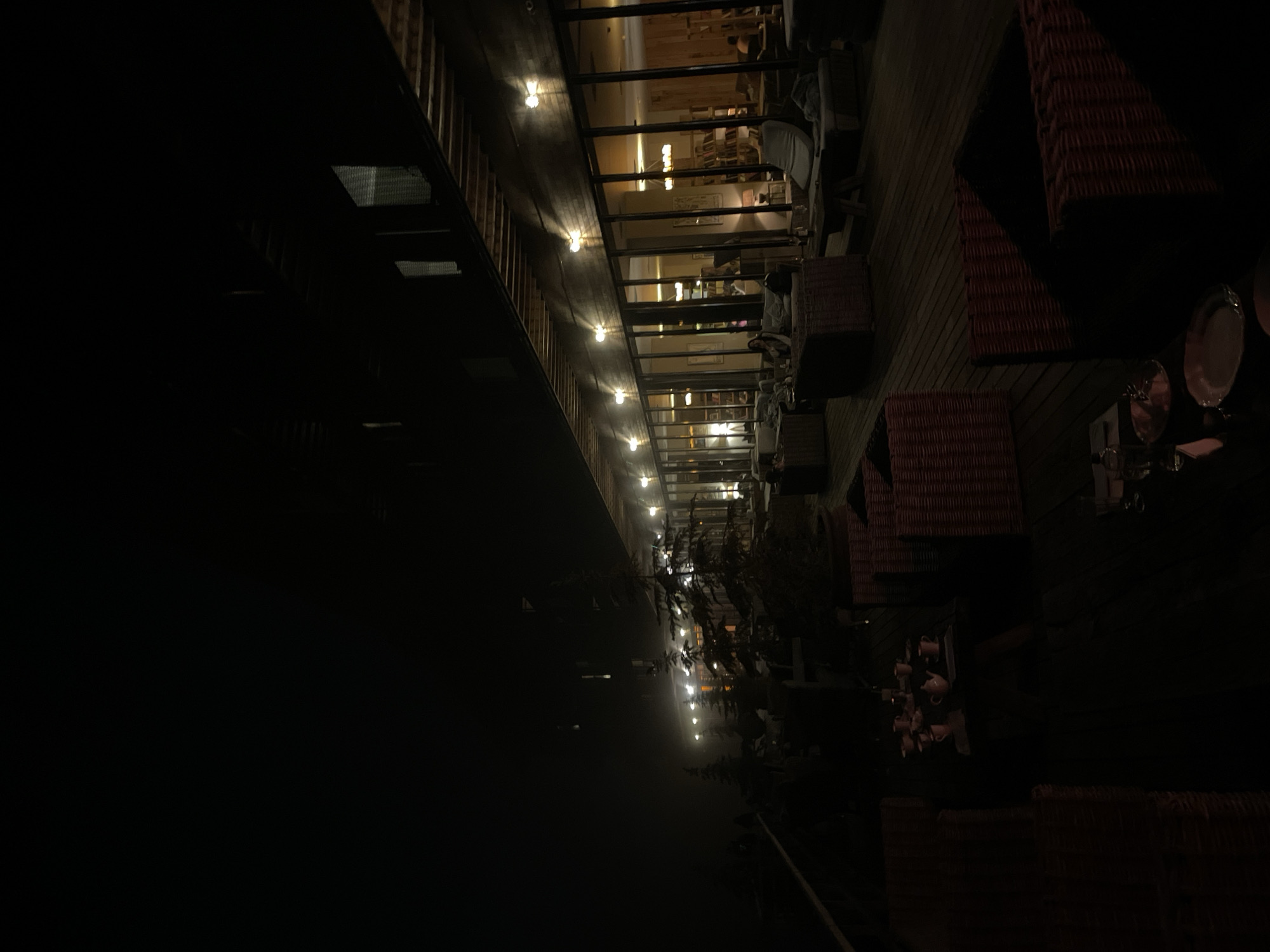 The image captures a dimly lit outdoor seating area of a restaurant or cafe at night, located in Stepantsminda, Georgia. The scene is characterized by a dark, moody atmosphere, with artificial lights casting a warm glow.
In the foreground, a wooden deck is visible, furnished with several wicker chairs and tables. On one table, a teapot and several cups are arranged, suggesting a recently served beverage. The chairs are predominantly dark, some with red accents, and are designed with intricate wicker weaving. Some of these chairs are empty, while others are occupied by patrons who are mostly obscured by the low light.
The middle ground reveals more seating areas and large glass windows that form the facade of a building. Through the windows, the interior of the establishment is visible, illuminated by warm lights and featuring bookshelves and lounge areas. Several individuals are seated inside, engaged in conversations or relaxing. Outside, a few trees, possibly pine or fir, are sparsely placed, their silhouettes visible against the dimly lit background. A faint mist or fog hangs in the air, contributing to the atmospheric effect.
The background shows the structure of the building, with a wooden exterior and a sloping roof. Outdoor lights are strategically placed along the building's edge, illuminating the deck and the surrounding area. The overall impression is one of a cozy and inviting, yet somewhat mysterious, nighttime setting.