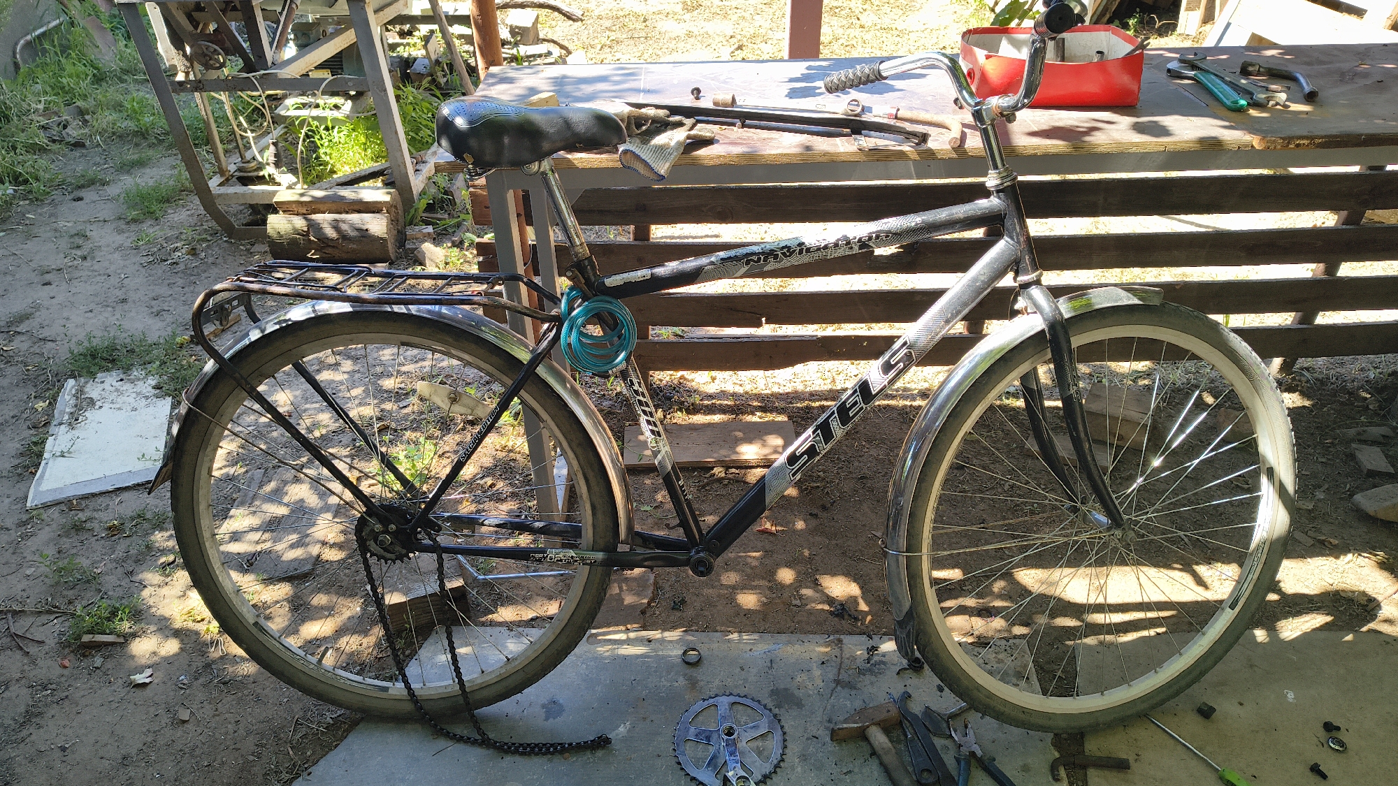 The image depicts a bicycle frame and wheels on a dirt and concrete surface, possibly an outdoor workshop or yard, in Mikhaylovka, Russia. A bicycle with a black frame, silver fenders, and a blue U-lock is partially visible. The front wheel is intact, while the rear wheel is detached and leaning. Various tools, including wrenches, pliers, and a hammer, are scattered on a wooden table and the ground nearby. A red toolbox sits on the table. The background shows greenery and what appears to be some sort of industrial equipment or structure. The lighting suggests it is daytime, with shadows cast by sunlight.