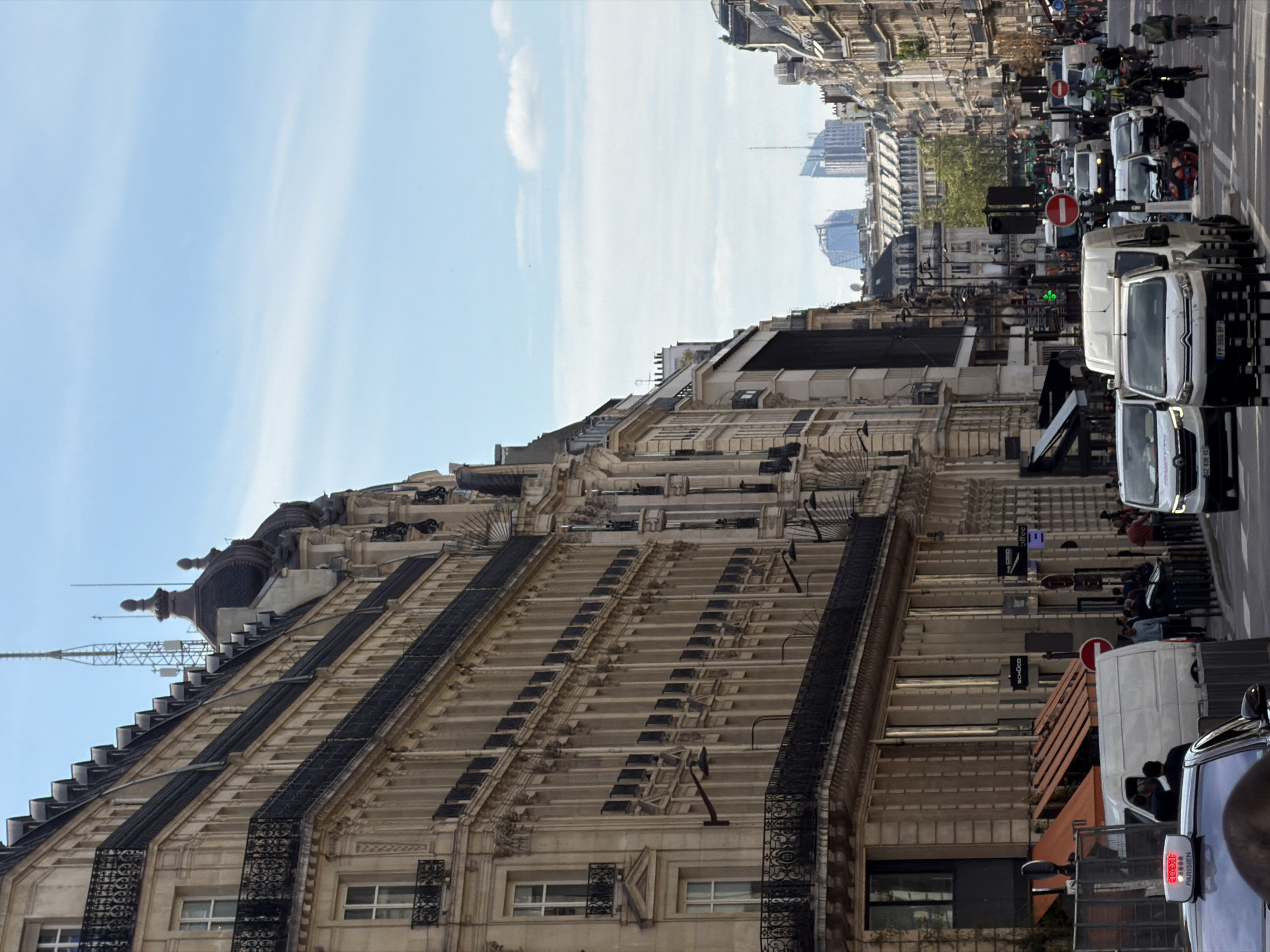 This photograph captures a street scene in Paris, France. The prominent architectural feature is a large, ornate building with multiple stories and detailed facades. It features wrought iron balconies and a distinctive roofline punctuated by finials and a lattice antenna. The building appears to be located on a busy street, where numerous vehicles, including vans, cars, and motorcycles, can be seen. Pedestrians are also present on the sidewalk. In the background, other buildings and skyscrapers create a dense urban skyline. The sky is partly cloudy.