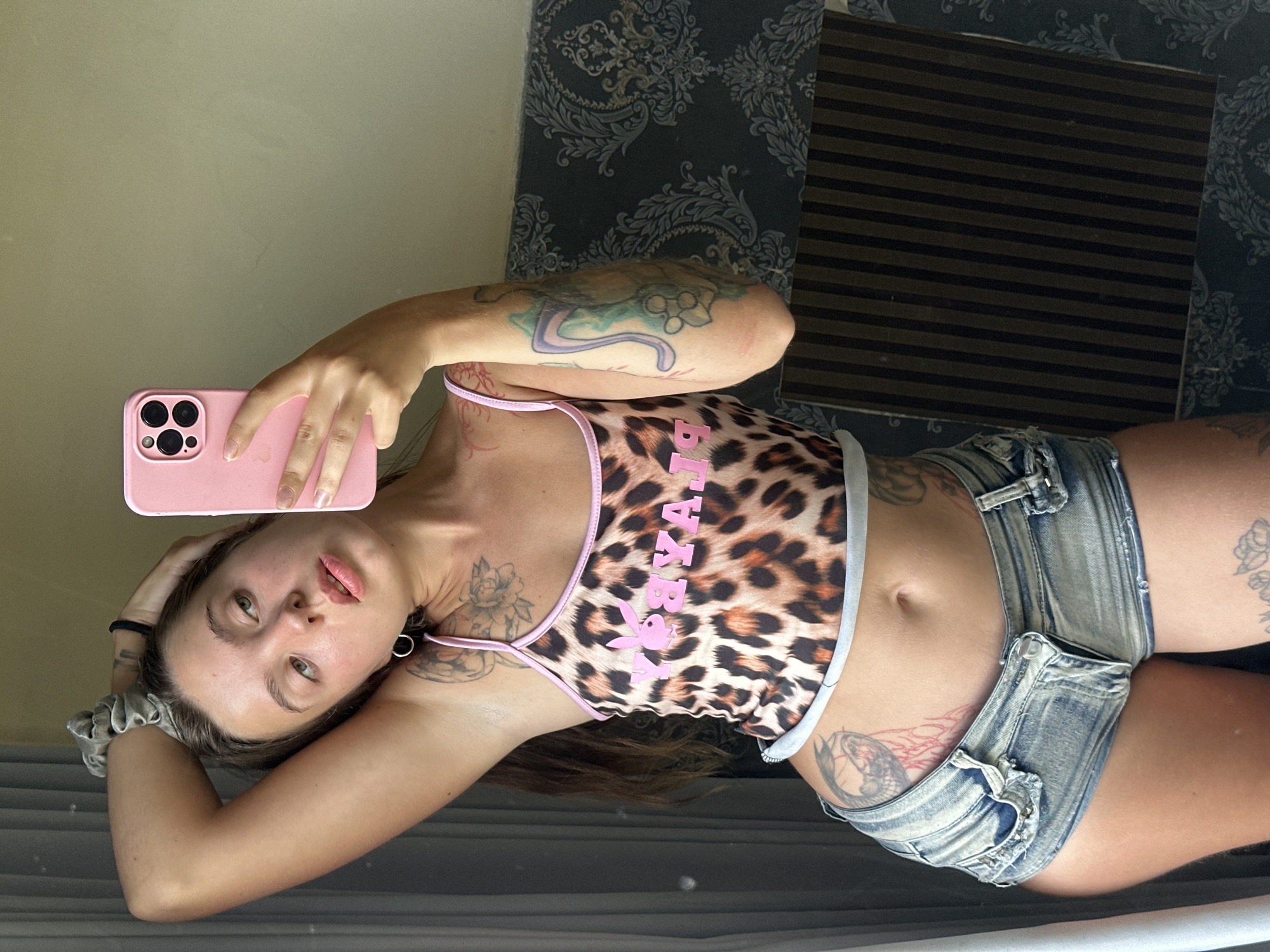 A young woman with long brown hair tied back with a grey scrunchie is taking a mirror selfie indoors. She is wearing a leopard print crop top with pink trim and the words "PLAYBOY" in pink lettering, and distressed denim short shorts. She has several tattoos visible on her arms, torso, and thighs. Her left arm is raised behind her head, and she is holding a pink smartphone in her right hand, angled to capture her reflection. The background consists of a sheer grey curtain on the left, a patterned wallpaper on the right, and a dark, vertically striped object in the lower right quadrant. The lighting suggests it is daytime.