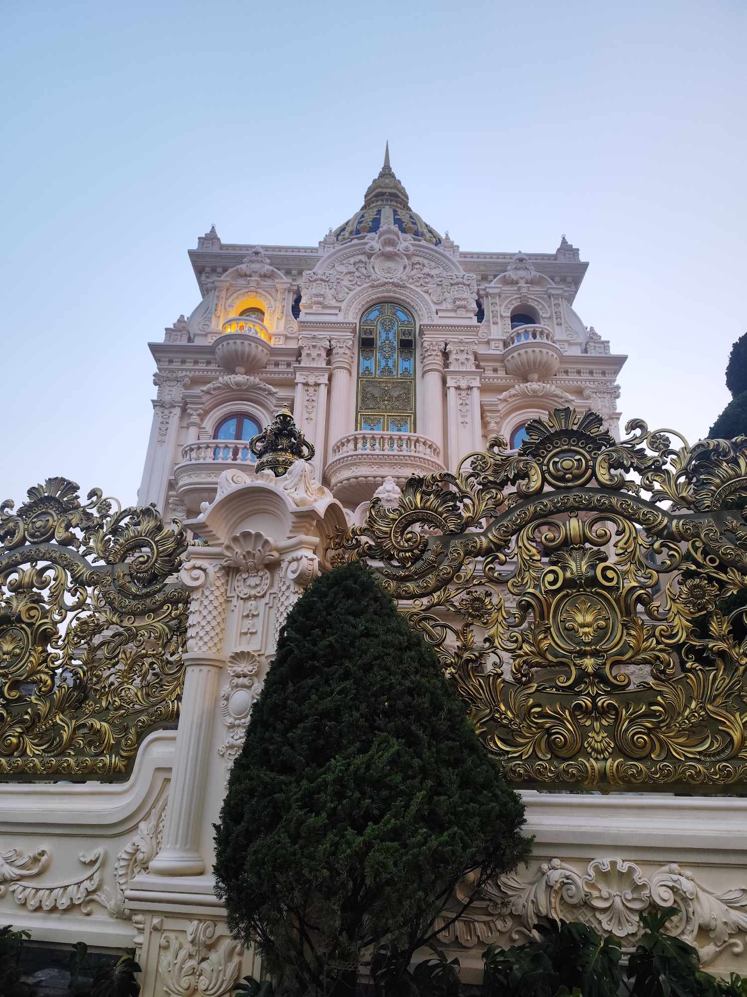 A low-angle shot depicts a large, ornate building, characterized by its elaborate architectural details and a towering presence. The structure is adorned with intricate carvings, ornate balconies, and a gilded wrought-iron fence at the base. The building's facade features a combination of pale stone and gold accents, creating a visually striking contrast. The sky above is a pale blue, suggesting either early morning or late evening. A conifer tree partially obscures the base of the building and fence. The photograph is taken at the Crazy House in Da Lat, Vietnam.