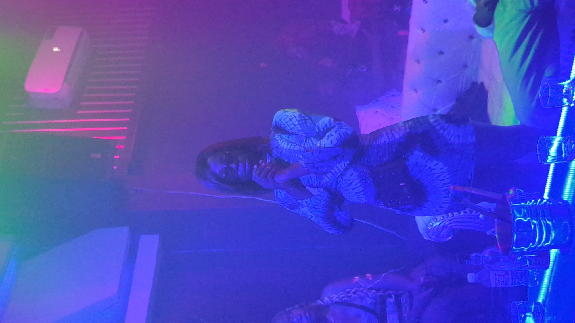 In Bar Jirgi Summa, Nigeria, a woman stands in a dimly lit establishment, illuminated by blue and pink lights. She wears sunglasses and a patterned dress, her hands clasped in front of her. To her left, another woman is partially visible, seated. In the foreground, a bar displays bottles of drinks, an ice bucket with tongs, and glasses. The background features a white tufted sofa and what appears to be an air conditioning unit on a wall with vertical accents. The atmosphere is smoky, contributing to the overall low-light conditions.