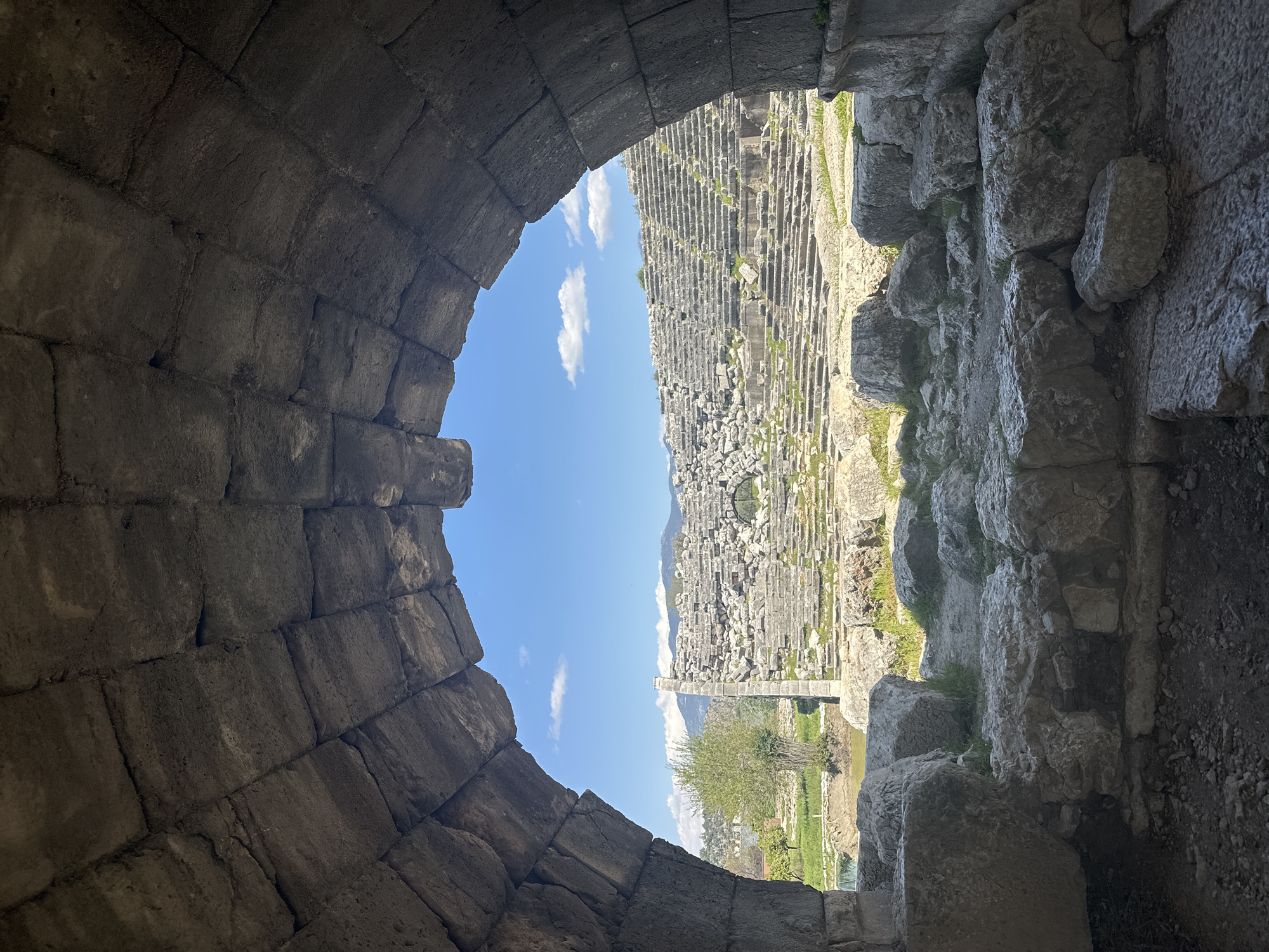 The image shows a view through an arched stone opening in Seydikemer, Turkey. The opening frames a scene of the ruins of an ancient amphitheater. The seating tiers of the amphitheater, constructed from stone, rise towards the sky. A single stone column stands within the ruins. In the background, below the amphitheater, is a small tree and a patch of green grass. The sky above is blue with a few scattered white clouds. The stone of the archway and the amphitheater shows signs of age and weathering.