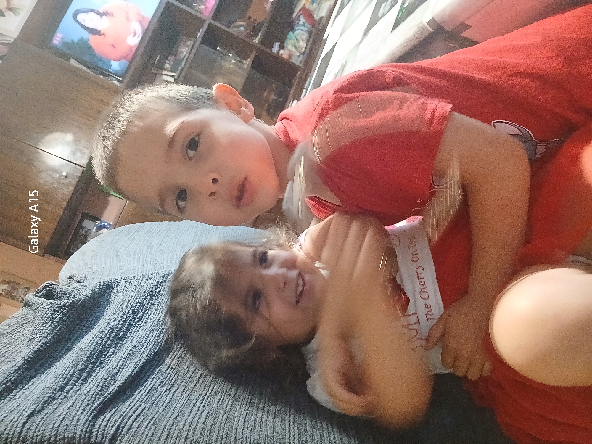 The image features two young children, a boy and a girl, engaged in a playful interaction on a bed or sofa in an indoor setting. The boy, wearing a red t-shirt, is in the foreground, lying on top of the girl and looking directly at the camera with a neutral expression. The girl, positioned beneath him and also wearing a red outfit with a white undershirt, smiles broadly towards the side with her arms raised, conveying a sense of joy or playfulness.
They are resting on a textured blue blanket or sheet. In the background, a brown wooden furniture piece, possibly a shelf, is visible. A television screen can be seen displaying what appears to be an orange cartoon character, possibly a fox.
Notable details include the text "Galaxy A15" written vertically along the left edge of the image, indicating the camera device. The girl's white undershirt prominently displays the phrase "The Cherry On Top." The lighting suggests an indoor scene, likely during the day or under artificial light. The overall mood is cheerful and relaxed. The location context for this scene is Obrochishte, Bulgaria.