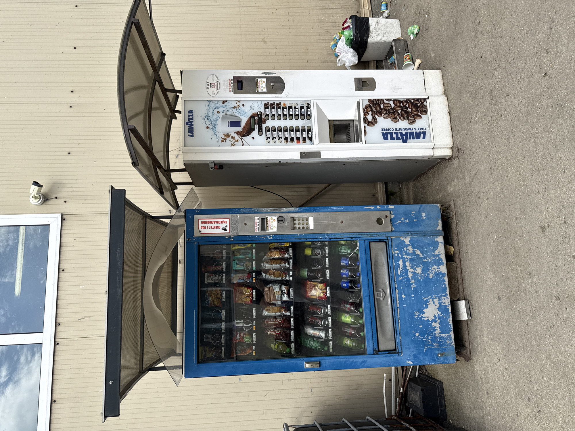 The image captures a street scene in Stara Zagora, Bulgaria, dominated by two vending machines. The blue vending machine at the bottom is for snacks, featuring various items like chips, chocolates, and beverages. Above it stands a coffee vending machine branded by Lavazza. The machines are positioned adjacent to a building with a window and a security camera mounted on its wall. Debris and litter are visible near the coffee machine, which is partially sheltered by an awning, indicating possible neglect or disuse. The ground appears to be a concrete surface, likely a sidewalk or pavement area. Text in Bulgarian is visible on the snack vending machine, suggesting its local origin and use.