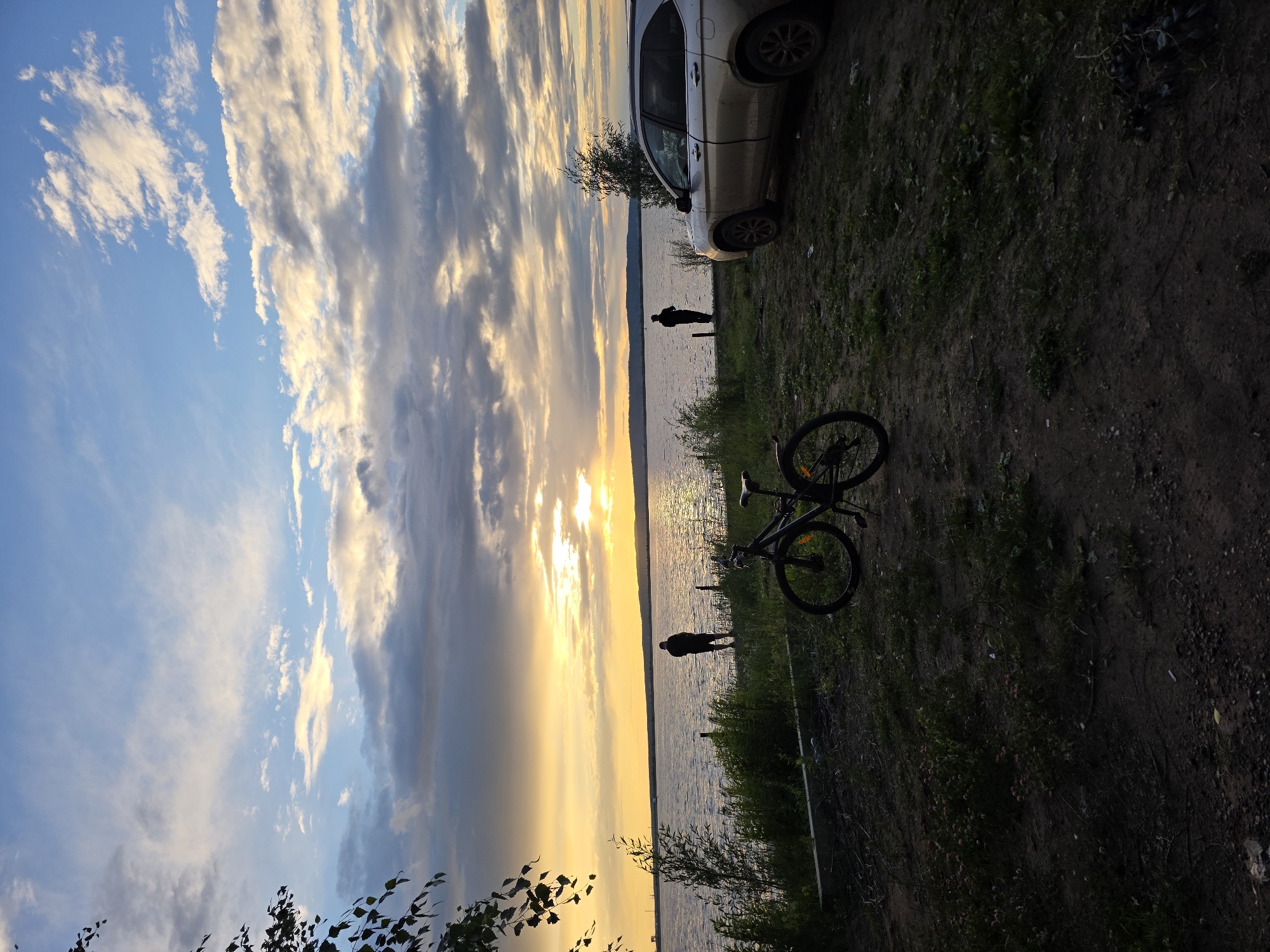 This image captures a serene sunset over a body of water, likely a river or lake, with a silhouette of a person standing near the water's edge in Chaikovsky, Russia. The sky is filled with dramatic, textured clouds, illuminated by the warm, golden hues of the setting sun.
In the foreground, a bicycle is parked on the grassy bank, suggesting a leisurely outing or a stop during a ride. Further back, near the water, another person is visible as a dark silhouette, seemingly contemplating the view. A car is parked on the bank behind the first person.
The scene evokes a sense of peace and tranquility. The time of day is clearly sunset, with the soft, warm light bathing the clouds and water. The weather appears calm, with no strong winds evident. The presence of a bicycle and people enjoying the natural beauty indicates a recreational setting, possibly a park or a designated spot for enjoying the outdoors. The overall mood is one of quiet reflection and appreciation for the natural landscape. There is no visible text in the image.