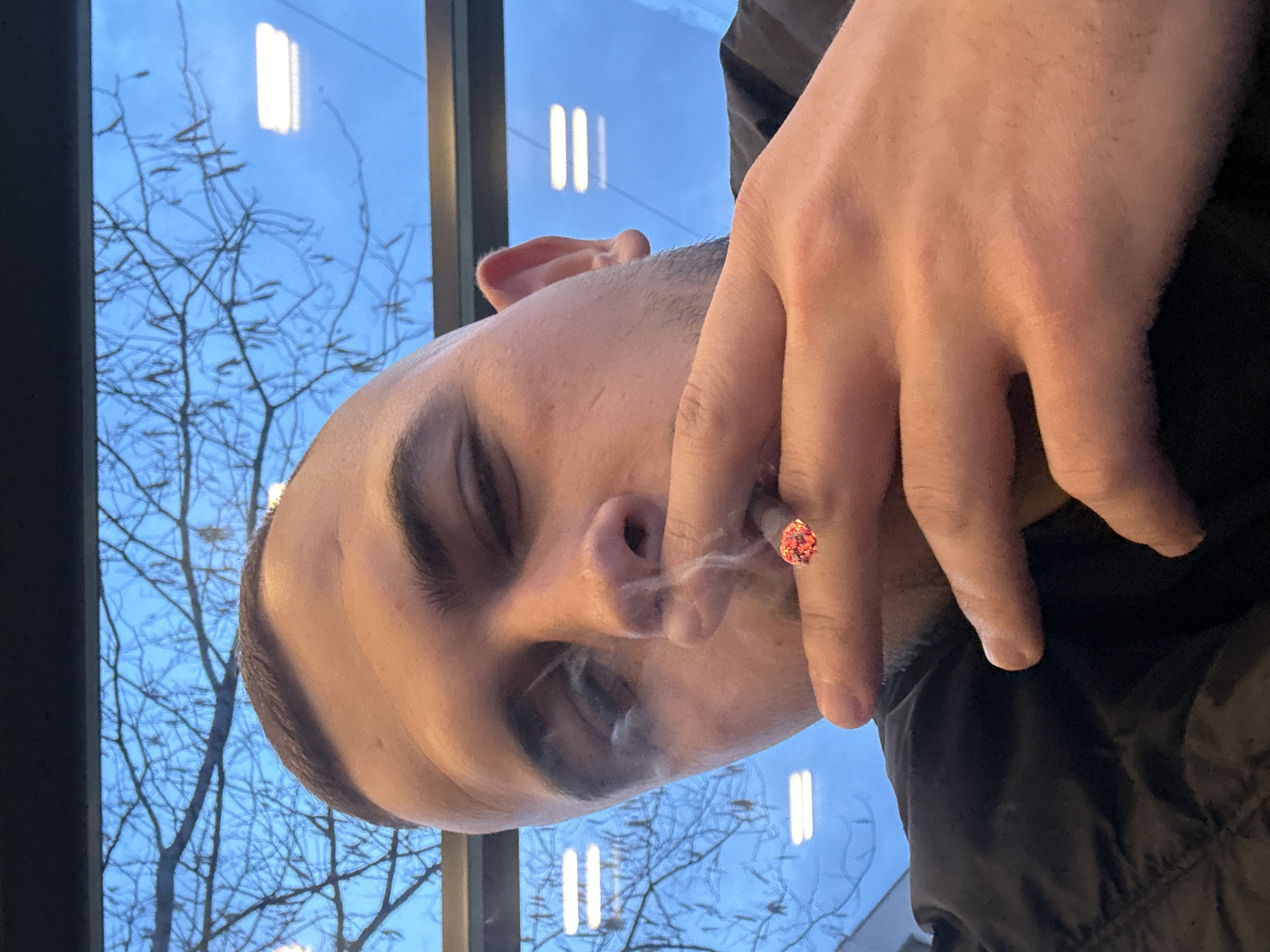 A close-up image features a male individual, approximately an adult, positioned in the foreground. He has short dark hair and visible stubble on his face. His right eye is partially closed, and his left eye is mostly obscured by the frame and smoke. The individual holds a lit cigarette between the index and middle fingers of his right hand, positioned near his mouth. The cigarette's tip glows orange-red, and white smoke emanates from it, partially obscuring his face. He is wearing a dark-colored, possibly quilted, jacket. The background, viewed through a window, shows a light blue sky. Bare tree branches, some with small catkin-like growths, are visible against the sky. Reflections of two sets of horizontal fluorescent strip lights are present on the window glass, indicating an indoor location. The visual elements within the image do not provide specific details to confirm its location as the city of Plovdiv, Bulgaria.