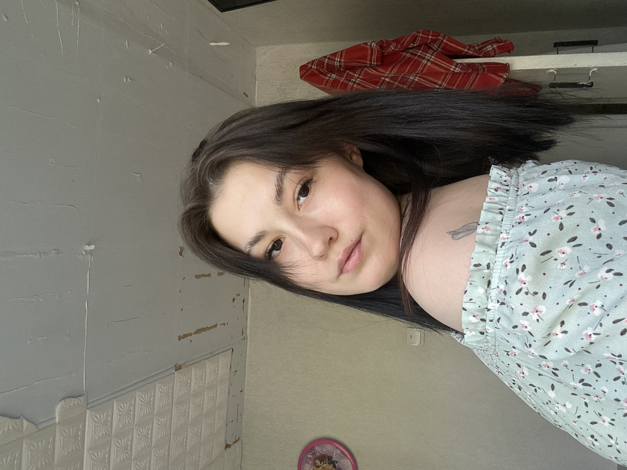 A young woman with dark hair is posing for a selfie in an interior setting, possibly in the city of Chaikovsky, Russia. She is wearing a light blue floral patterned dress with a ruffled neckline. A portion of a tattoo is visible on her shoulder. Behind her, there is a textured grey wall with decorative molding. Above the wall, there's what appears to be a cabinet or shelf with a red plaid garment on top. On the floor, partially visible, is a round, pink decorative item, possibly a clock or ornament.