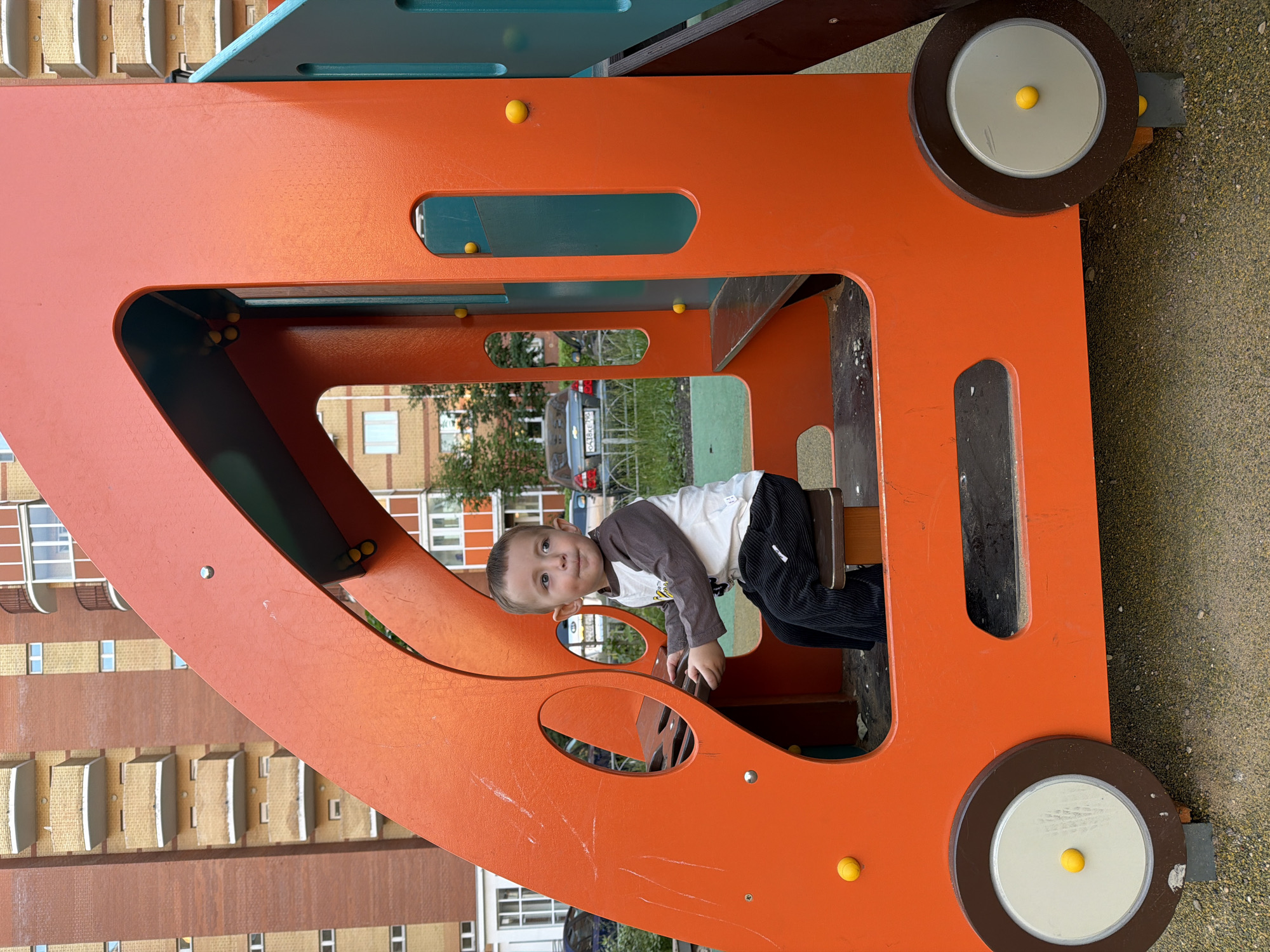A young Caucasian boy with short brown hair and light skin is sitting inside a bright orange children's playground structure designed to resemble a vehicle. He is wearing a long-sleeved shirt with a white body and brown sleeves, and dark corduroy pants. The boy is holding onto a steering wheel-like element, looking to his right with a slightly open mouth. The playground structure features yellow spherical decorative elements and a gray seat. In the background, there is a tall, multi-story apartment building with brick and yellow facade, and balconies. Several cars are parked near the building. The ground is covered with small pebbles and sand. The setting appears to be a residential courtyard in the city of Vniissok, Russia.