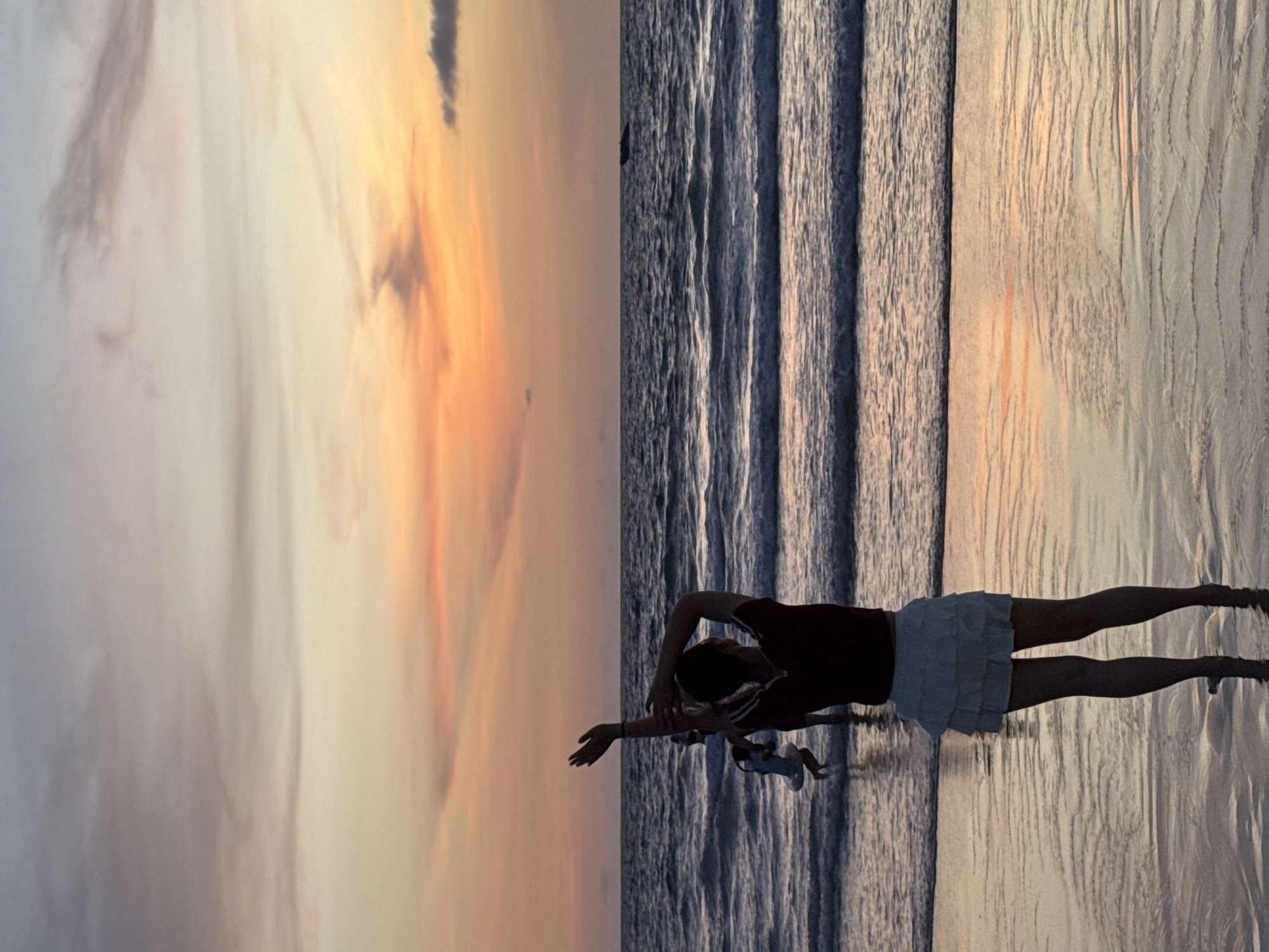 A full shot captures a beach scene at sunset, likely in Karon, Thailand. In the foreground, a person stands facing away from the camera, with their arms raised and hands reaching upwards, suggesting a pose of freedom or enjoyment. They are wearing a dark red top and a white, ruffled skirt. In the middle ground, a child is visible in the water, and further out in the ocean, a boat is discernible. The sky is painted with hues of orange, pink, and yellow from the setting sun, casting a warm glow on the scene. Gentle waves roll onto the shore, creating a serene and picturesque atmosphere.