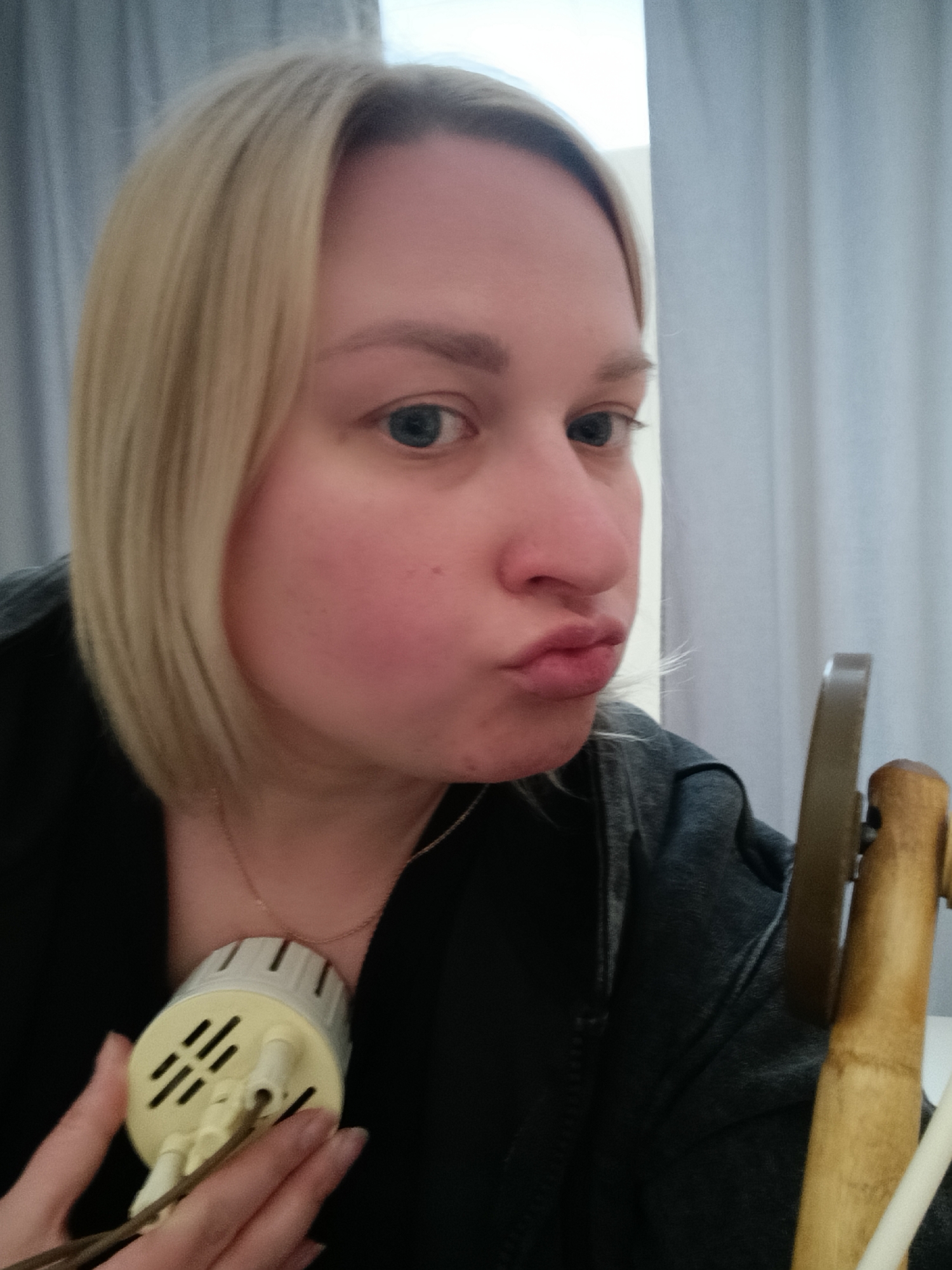 A fair-skinned woman with short blonde hair and blue eyes is holding a small, cylindrical, off-white electrical device with slots and prongs in her left hand near her chest. She is wearing a black jacket and a thin gold necklace. Her lips are pursed in a kiss-like expression. In the background, there is a light-colored curtain. To the right, parts of a light-colored, possibly bamboo-like structure are visible. The location is Chaikovsky, Russia.