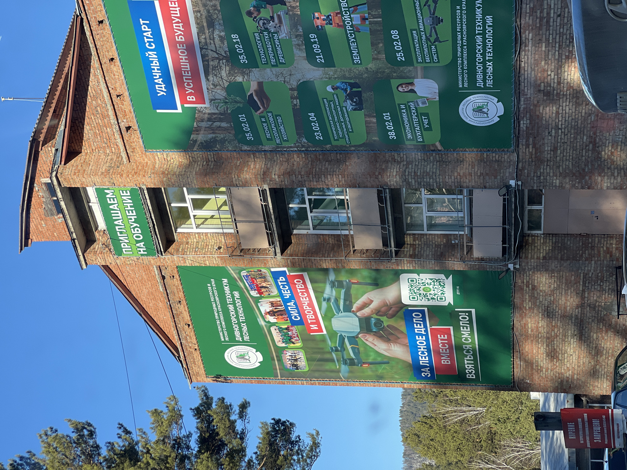 The image shows a red brick building in a city (location unknown) adorned with large green banners. The banners promote Divnogorsk Forestry College, featuring various specializations such as forestry, woodworking technology, land management, drone operation, and accounting. One banner displays images of students and a drone, with slogans like "Power, Honor, and Creativity" and "Together for the forestry business! Let's be brave!". A sign reading "Smoking is prohibited" is visible in the foreground, indicating a non-smoking policy in the area.
