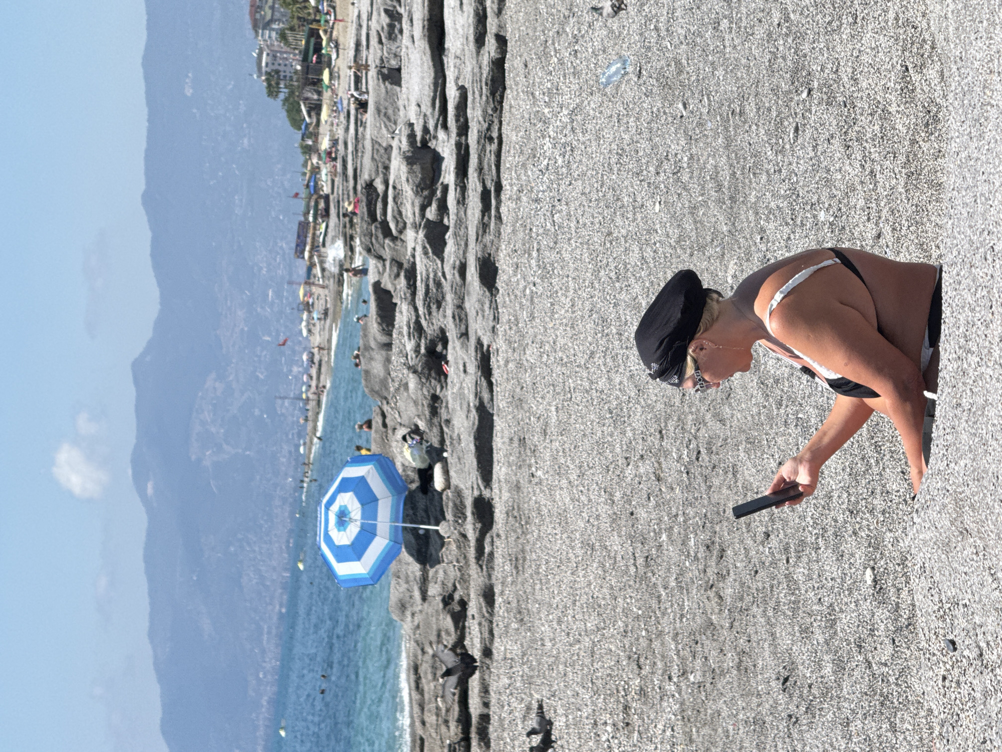 The image depicts a beach scene. A woman in a bikini and a black cap is bending over, holding a black object, possibly a phone. The beach is sandy, with pebbles scattered around. Behind the woman, there's a rocky cliff with people and umbrellas scattered on it. Further in the background, there's the sea, with a blue and white umbrella near the shore. In the distance, there is a city or town, and a mountain range is visible on the horizon under a clear sky. The location appears to be a beach resort. The city is Unknown, Unknown.