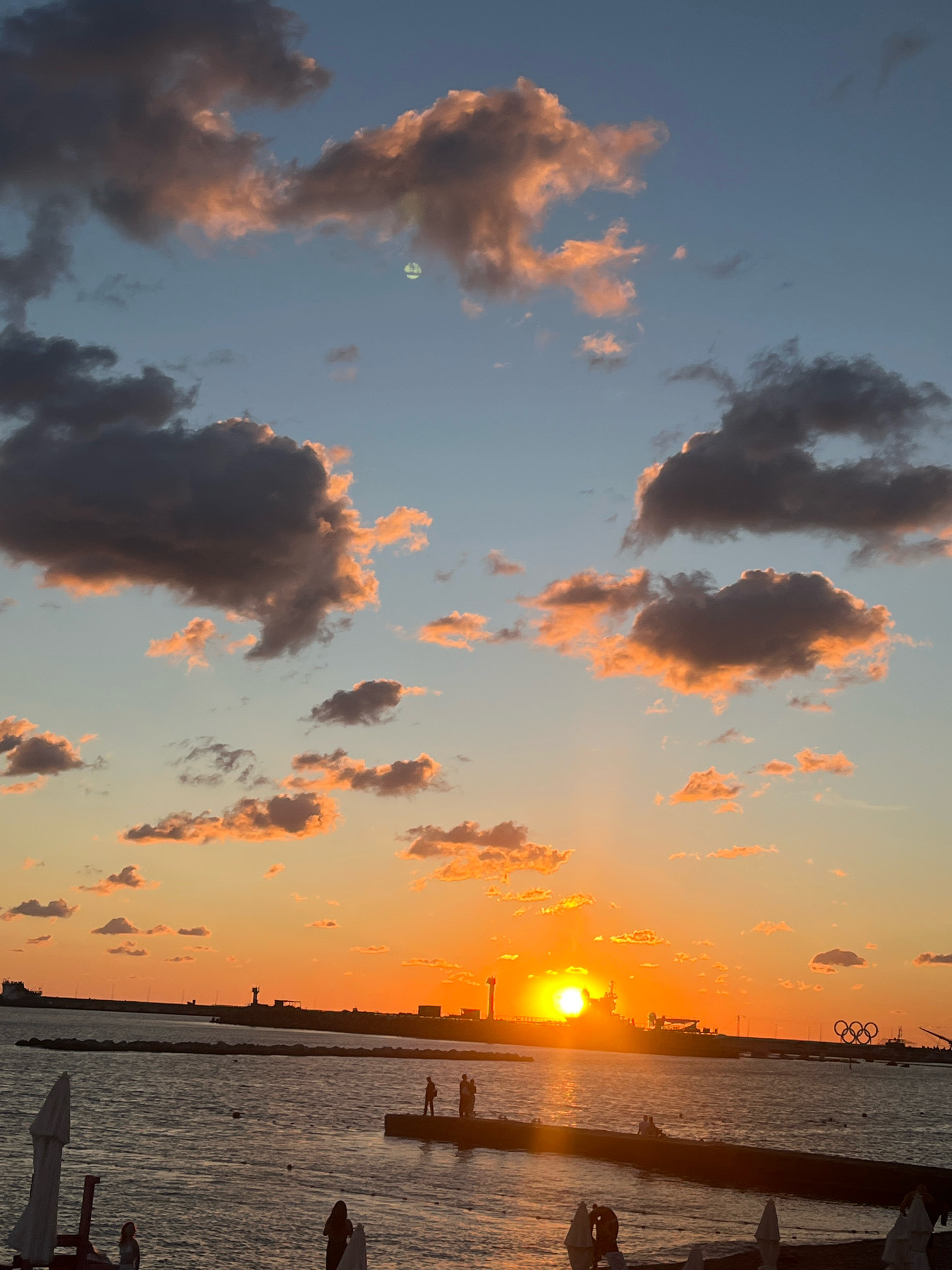 A wide-angle, vertical, outdoor, daytime, medium shot captures a sunset over the Black Sea in Sochi, Russia. The sky is a gradient of soft blues and oranges, with scattered clouds tinged pink and yellow by the setting sun. The sun itself is a bright, radiant orb low on the horizon, casting a golden glow across the water. Silhouettes of people are visible on a pier in the foreground, enjoying the view. A long, dark breakwater extends into the sea, with industrial structures, including a lighthouse, visible in the distance. Olympic rings are subtly present on the right side of the frame. The water is calm, reflecting the colors of the sky. Umbrellas and beach chairs are visible on the shore in the foreground, suggesting a public beach area.