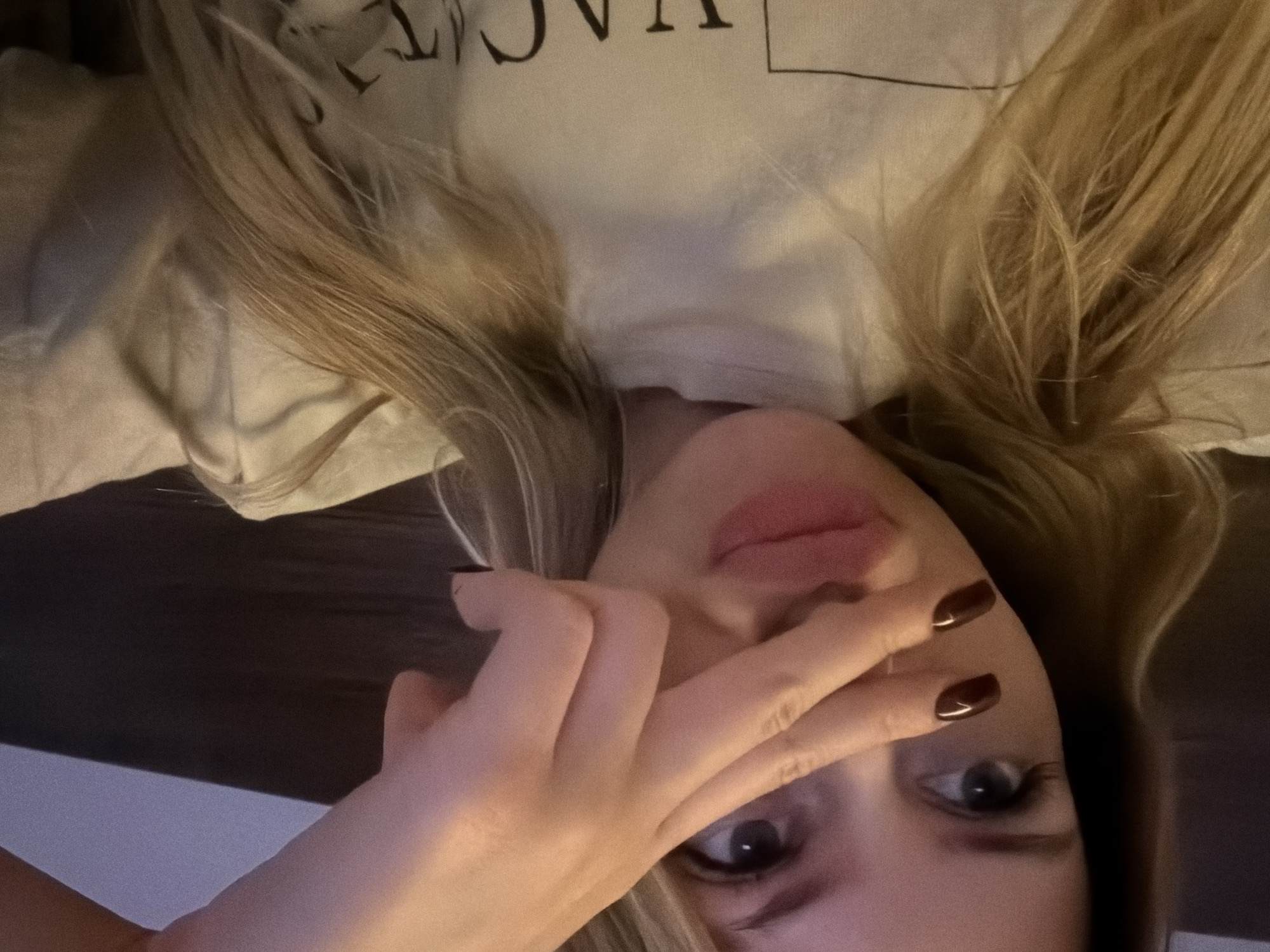 A close-up, selfie-style shot shows a young Caucasian woman with blonde hair and fair skin. She is wearing a light-colored t-shirt with a partially visible graphic print. Her right hand, with dark nail polish, is held up to her face, obscuring her nose and mouth. Her left eye is visible, with apparent eyelashes. The background is dark and out of focus, with a hint of a patterned surface. The lighting is dim, creating a soft, warm glow. There is no identifiable information about the location within the city of Đà Nẵng, Vietnam.