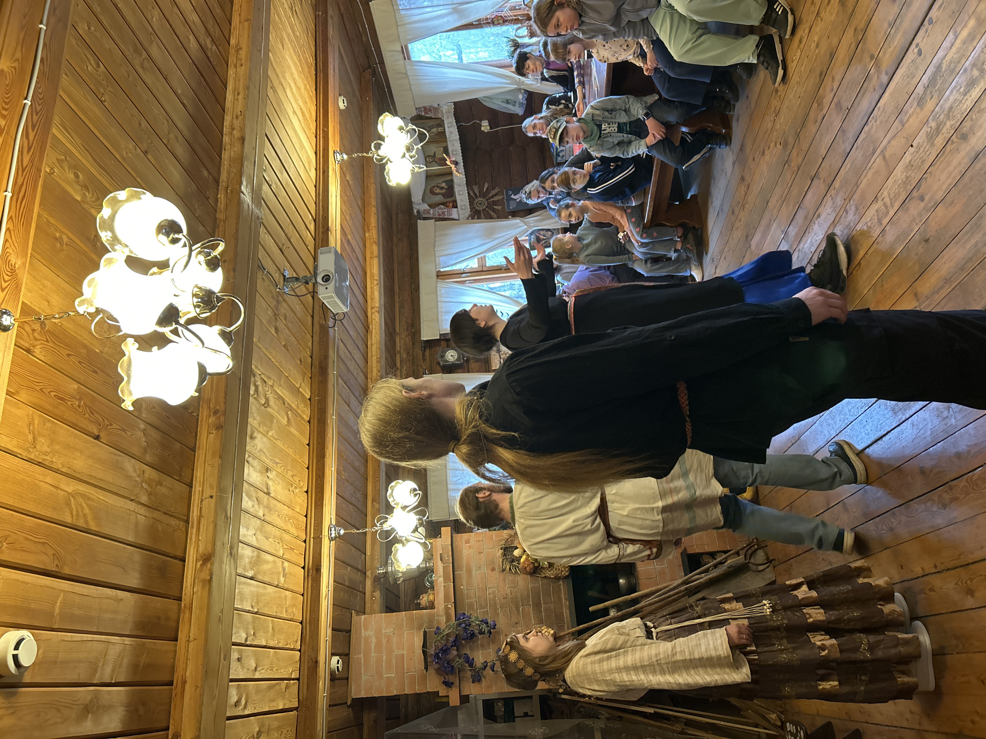A group of people are gathered in a wood-paneled room with hardwood floors. The room has a rustic, possibly historical, aesthetic, with exposed wooden beams and period-style chandeliers. A projector is mounted on the wooden wall.
Several people are seated in the background, facing a woman in a black robe who is gesturing with her hands as if speaking or giving instructions.
In the foreground, three people are standing. One has long blond hair pulled back into a ponytail. Below them is a brick fireplace with decorations.
At the very front is a person dressed in what appears to be a historical or theatrical costume, possibly portraying a character or figure from a historical event. This person is wearing a wreath on their head and holding sticks or staffs.