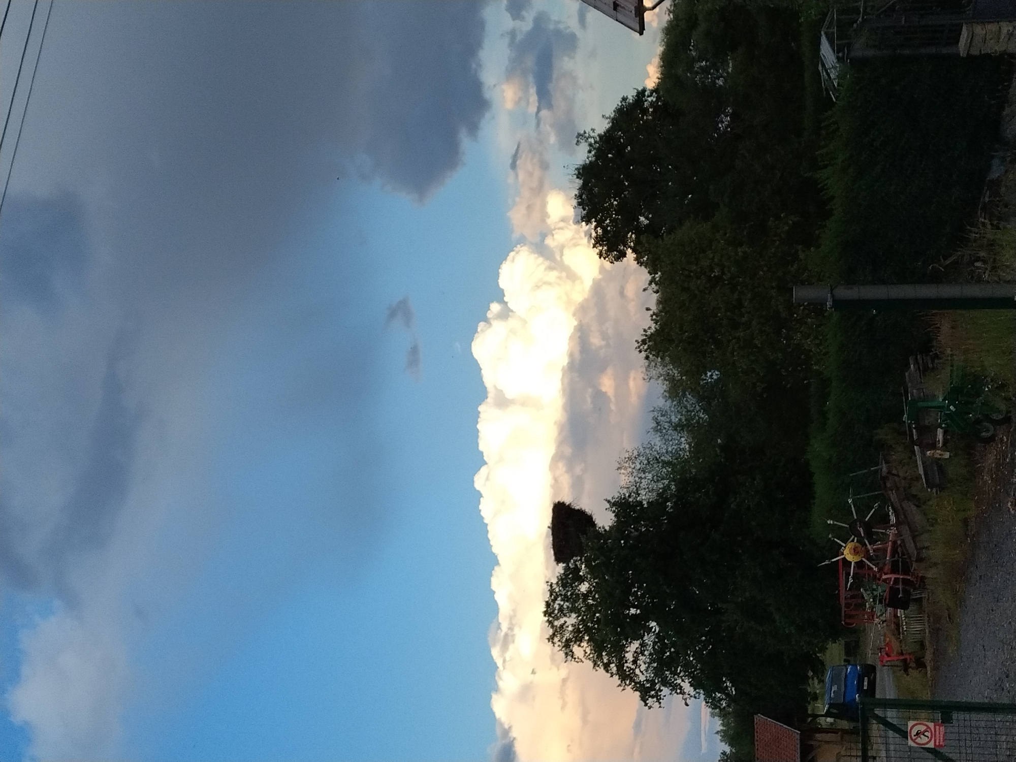 The image depicts a rural scene in Dobev, Czechia. In the upper portion of the frame, a dramatic sky features both bright blue expanses and heavy, dark clouds, with a large, luminous cloud bank prominently displayed. A stork's nest, perched atop a mature tree, is visible against this backdrop. Below the sky, a dense line of dark green trees forms a natural barrier. In the foreground, a gravel driveway leads past a metal gate with a "no trespassing" sign. To the left, a portion of a building with a red-tiled roof is visible. Further into the scene, various pieces of agricultural machinery are parked, including a blue van, a red trailer-like structure, and other implements, some of which appear weathered and overgrown with grass. A dark green hedge runs horizontally across the middle ground, with a small greenhouse partially obscured behind it on the right. The overall impression is one of a working farm or rural property in the late afternoon or early evening.