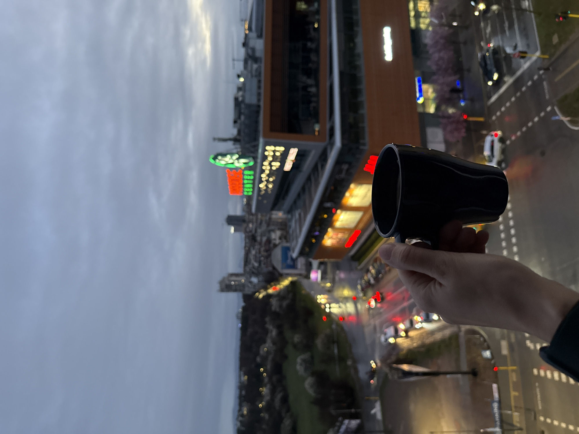 The image shows a nighttime cityscape in Sofia, Bulgaria, viewed from an elevated position, possibly a balcony or high-rise building. A hand is holding a black cylindrical object in the foreground. The city is visible in the background, including buildings with bright lights and neon signs in Cyrillic characters. Streetlights and car headlights illuminate the roads and sidewalks. The sky is overcast, with a gray-blue hue.