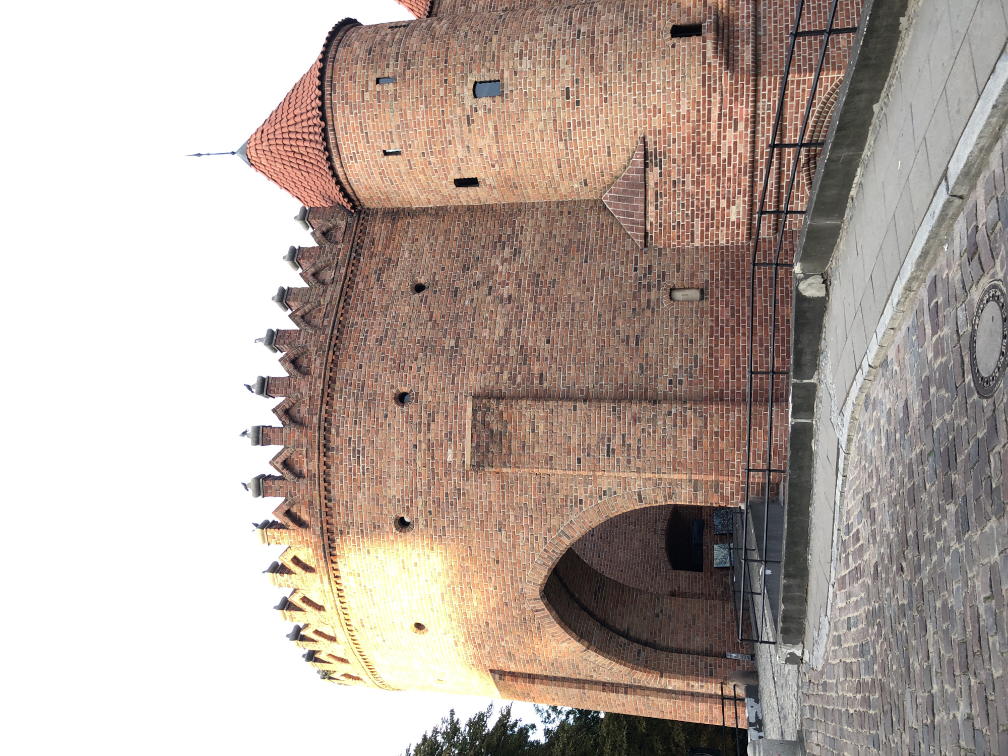 This is a full-shot, eye-level, outdoor, daytime photograph of the Barbican in Warsaw, Poland. The Barbican is a historic fortification made of red brick, with a large arched entrance on the left side of the frame. A conical red-tiled tower with a pointed spire rises from the right side of the building. The top of the Barbican is adorned with battlements, on which several pigeons are perched. A pathway made of cobblestones leads up to the Barbican, and a gray paved walkway with a black metal railing runs alongside it. A few trees are visible on the far left, and the sky is clear and white. The lighting suggests it is late afternoon or early morning, as the sun casts a warm glow on the brickwork.