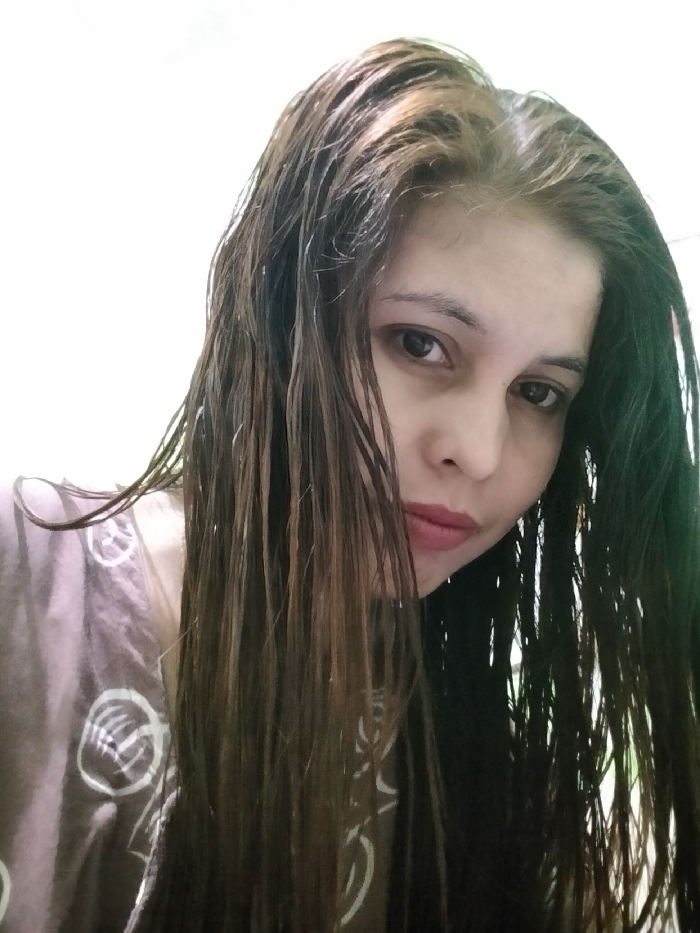 The image shows a female subject captured in a close-up portrait, reportedly taken in an indoor environment in Ranugedang, Indonesia. She is positioned from the chest up, facing the camera with a direct gaze. Her long, dark brown hair, appearing wet or damp, cascades over her shoulders and chest, partially obscuring the sides of her face. Lighter brown highlights are visible, particularly near the crown. The subject has light skin, dark eyes, and her lips are closed, colored with a reddish-pink hue. She is wearing a light brown or mauve garment featuring white circular patterns visible on the left side of the frame. The background is a plain, uniformly lit, light-colored surface, likely white or off-white. The subject is observed posing for a photograph.