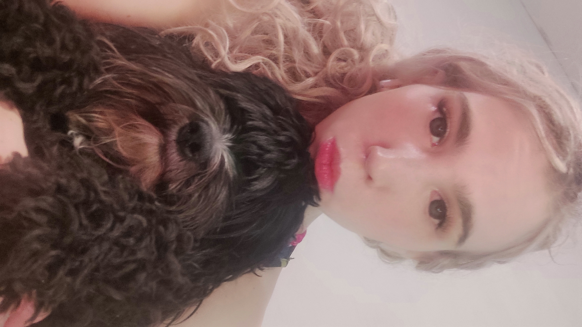 A fair-skinned young woman with light blonde curly hair holds a small, dark-haired dog close to her face. The woman has fair skin, light eyebrows, and pink lips. The dog has a shaggy coat and brown highlights around its muzzle. The background is a blurred white. The image appears to be taken indoors, possibly in a residential setting. The location in the city of Chaikovsky, Russia is indeterminable from the image.