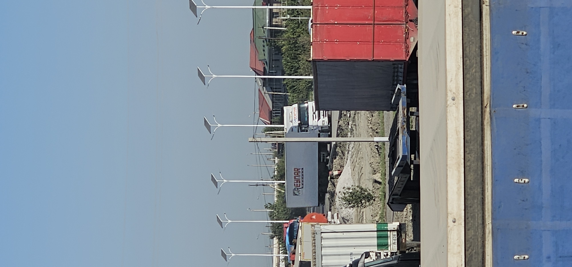 The image shows a street scene in Sheyhancha, Uzbekistan, with several large trucks parked and moving. In the foreground, there is a large blue structure with metal fixtures, possibly part of a building or a large vehicle. Behind this, a flatbed trailer is visible. Further back, a red truck is parked next to a grey truck. To the left, a white truck with green and white stripes is parked. In the middle ground, a white semi-trailer truck with "GREYNAR" written on its side is visible. In the background, there are more trucks, solar-powered streetlights, houses with red and green roofs, and trees. The sky is clear and blue.