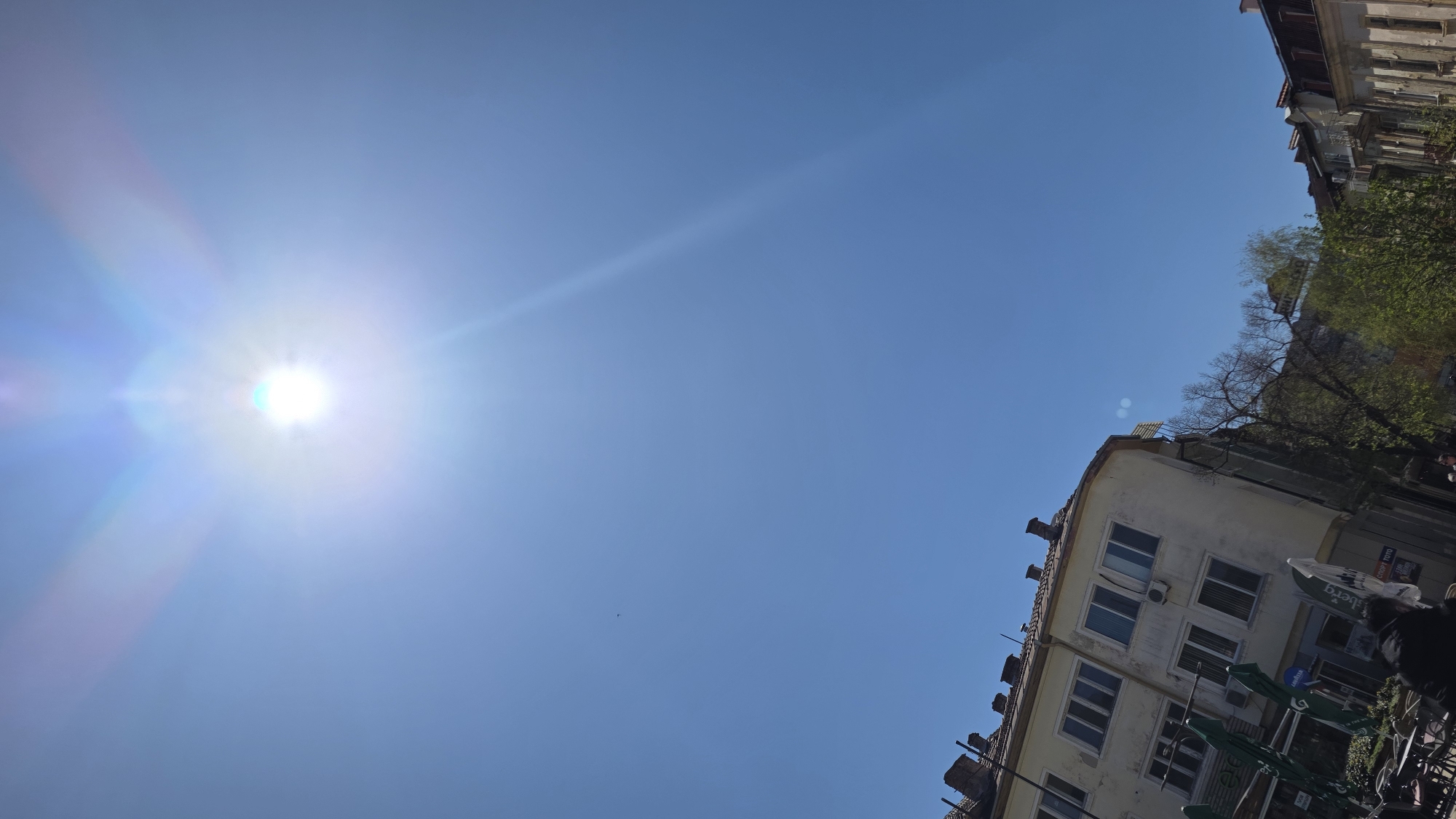 The image depicts a bright, sunny day in what appears to be an urban setting. The sun is prominent in the upper left of the frame, causing significant lens flare. A clear blue sky dominates the majority of the image. In the lower right corner, a building with multiple windows and a light-colored facade is partially visible. Green trees are present above the building. Tables with umbrellas are positioned in front of the building on the ground. Based on the visible Berg logo, it is likely that this location is in Blagoevgrad, Bulgaria.