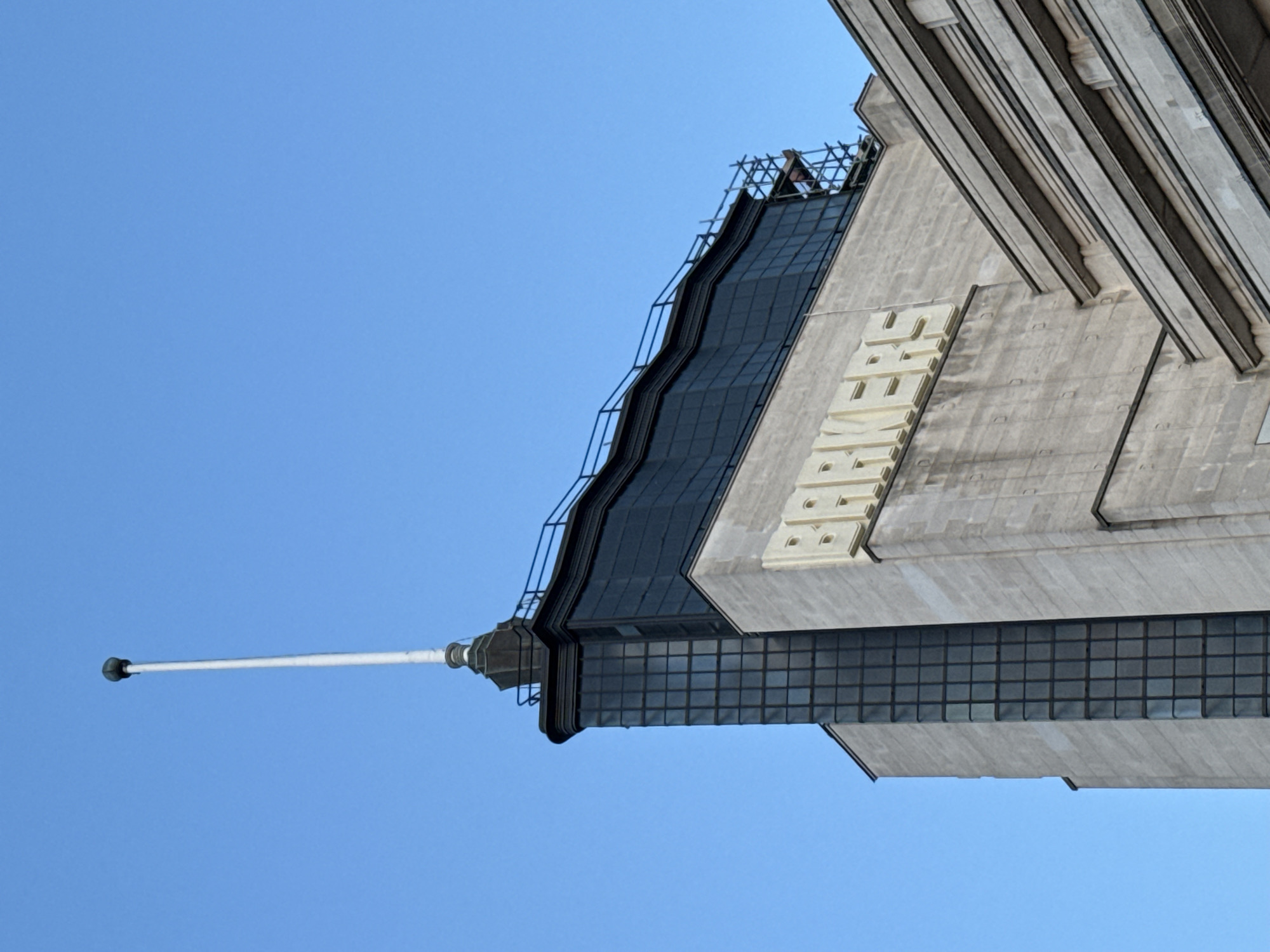 The image shows the top portion of a building against a clear blue sky. The building's architectural style appears to be Art Deco, with a prominent spire and decorative elements. A white rod, likely a lightning rod, extends from the top of the spire into the sky. The building's facade features a grid of windows and stone or concrete ornamentation. In the lower part of the image, a series of vertical ribs or columns are visible. The building is located in the City of London, United Kingdom. The name of the building is engraved in large letters on the side.