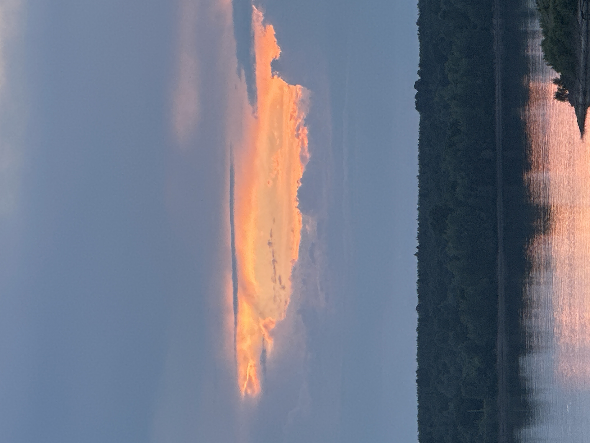 The image depicts a tranquil body of water, likely a river, at dusk. The sky is a gradient of soft blues and grays, with a prominent band of orange and pink clouds illuminated by the setting sun. A dense, dark green forest lines the opposite bank of the water, forming a stark contrast to the colorful sky. The water reflects the colors of the sky and the trees, creating a mirrored image of the scene. The overall impression is one of natural beauty and serenity. The scene is consistent with views along the Kama River in Chaikovsky, Russia, particularly at sunset. There are no people or artificial structures visible in the image.