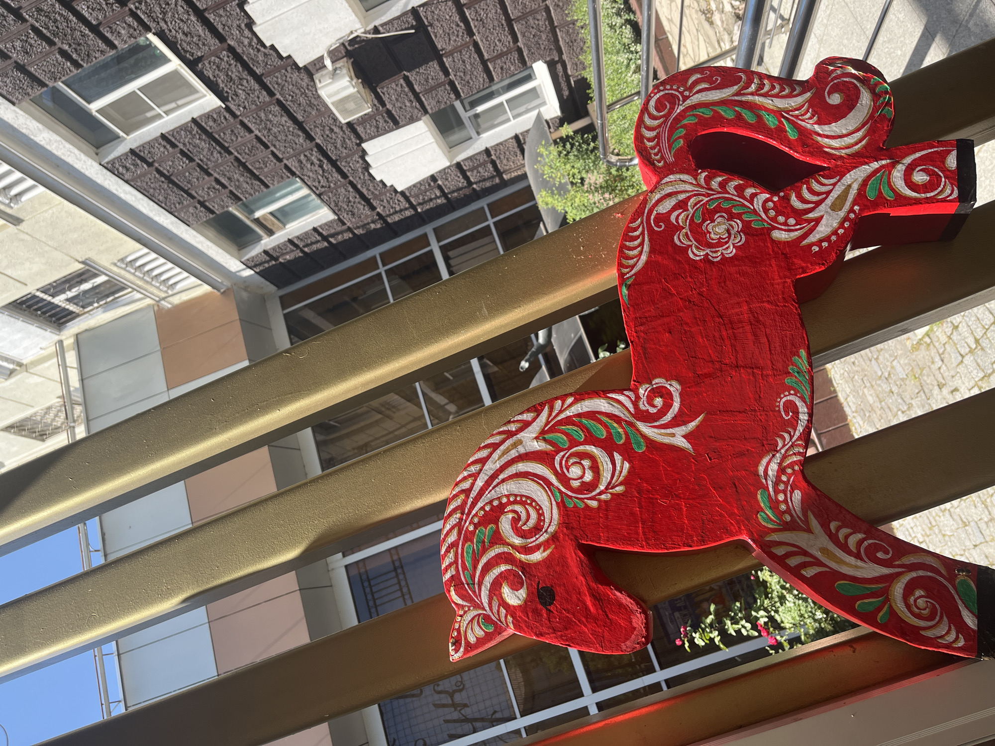 A red, wooden, decorative horse with white and green folk-style patterns is positioned outdoors in front of a building. The horse is in the foreground and is partially obscured by gold-colored vertical beams. In the background, there is a multi-story building with a dark, textured facade and windows. Green bushes and a metal railing are also visible. The sky is blue and clear. The scene appears to be located in a city environment, possibly in Krasnodar, Russia, based on the architectural style. There are no people visible in the image.