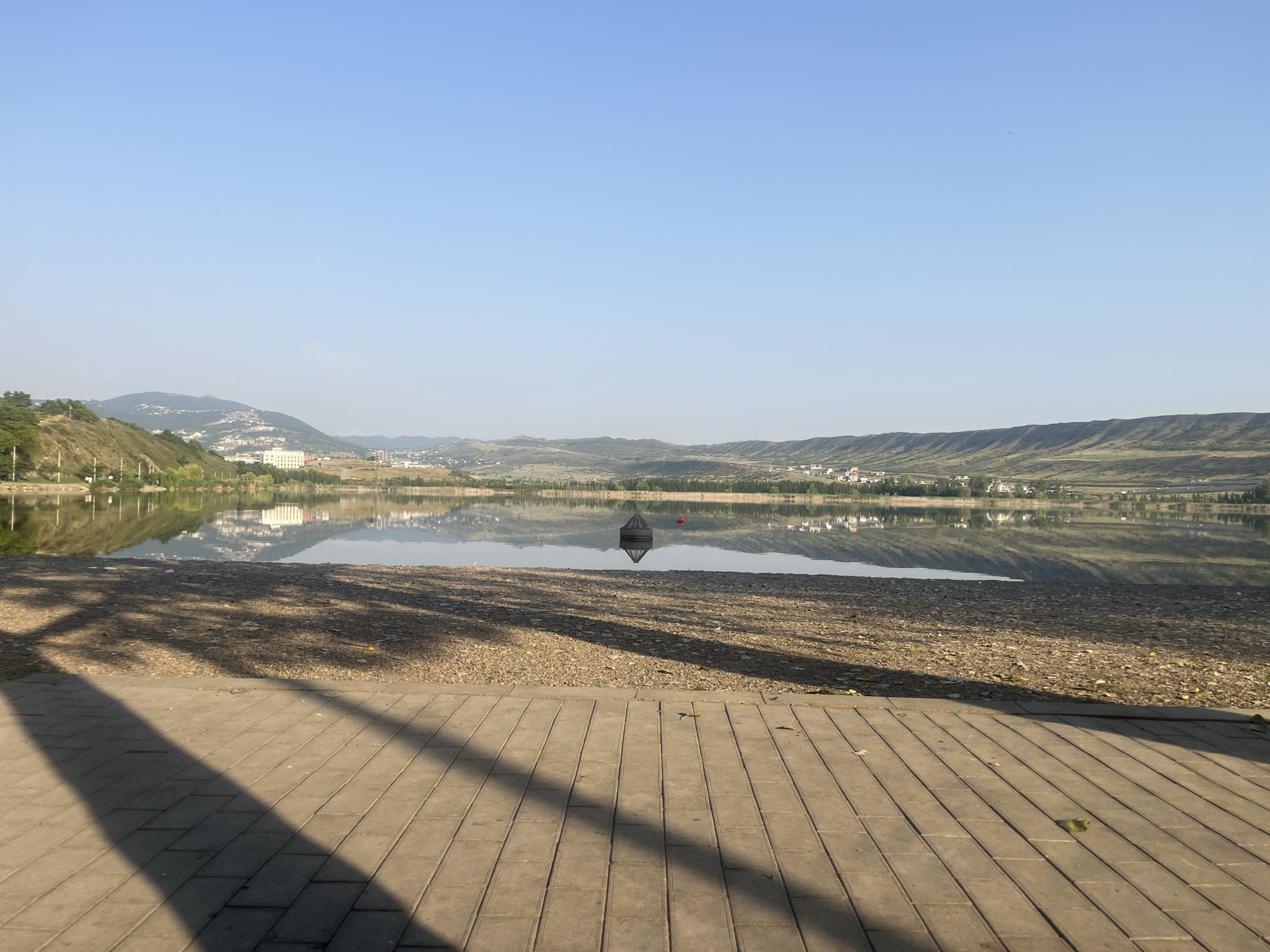 The image shows a serene lake scene with a paved walkway in the foreground. The walkway is made of rectangular concrete tiles laid horizontally, with a shadow of what appears to be a tree cast across it diagonally from the left. Beyond the walkway is a beach area covered in small pebbles and dirt.
The lake itself is calm, with clear reflections of the distant landscape. In the middle of the lake, there is a dark, pyramidal structure that seems to be a buoy or a decorative element, with a red spherical buoy nearby. The far side of the lake is lined with rolling hills covered in green vegetation and scattered buildings. The architecture of the buildings suggests a semi-urban or suburban setting, with some larger structures visible on the hillsides. The sky is clear and blue, indicating a bright, sunny day.
This scene is likely the Tbilisi Sea (also known as the Tbilisi Reservoir), a large artificial lake located on the northeastern outskirts of Tbilisi, Georgia. It serves as a major water source for the city and is a popular recreational area. The presence of the paved walkway and beach area indicates a developed public space designed for visitors. The distant hills and the architectural style of the buildings are consistent with the geography and development patterns around Tbilisi.