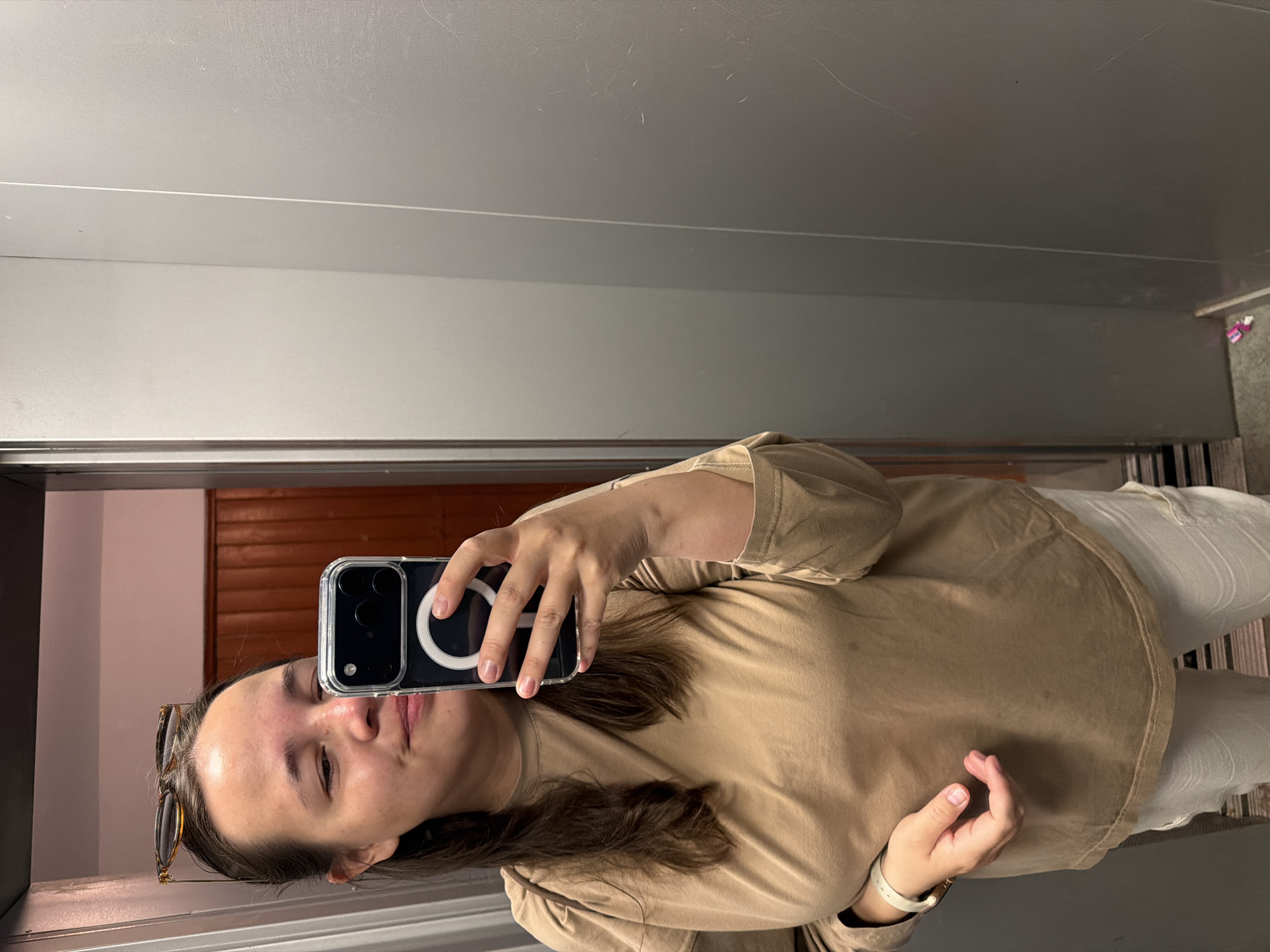 A person with brown hair, wearing a beige shirt and white pants, is taking a selfie in a bathroom. The person is holding a phone with a clear case and a MagSafe ring on the back. There are sunglasses resting on their head. The bathroom has grey walls. Through the mirror, a section of a door or wall with brown horizontal panels is visible. The location is Chaikovsky, Russia.
