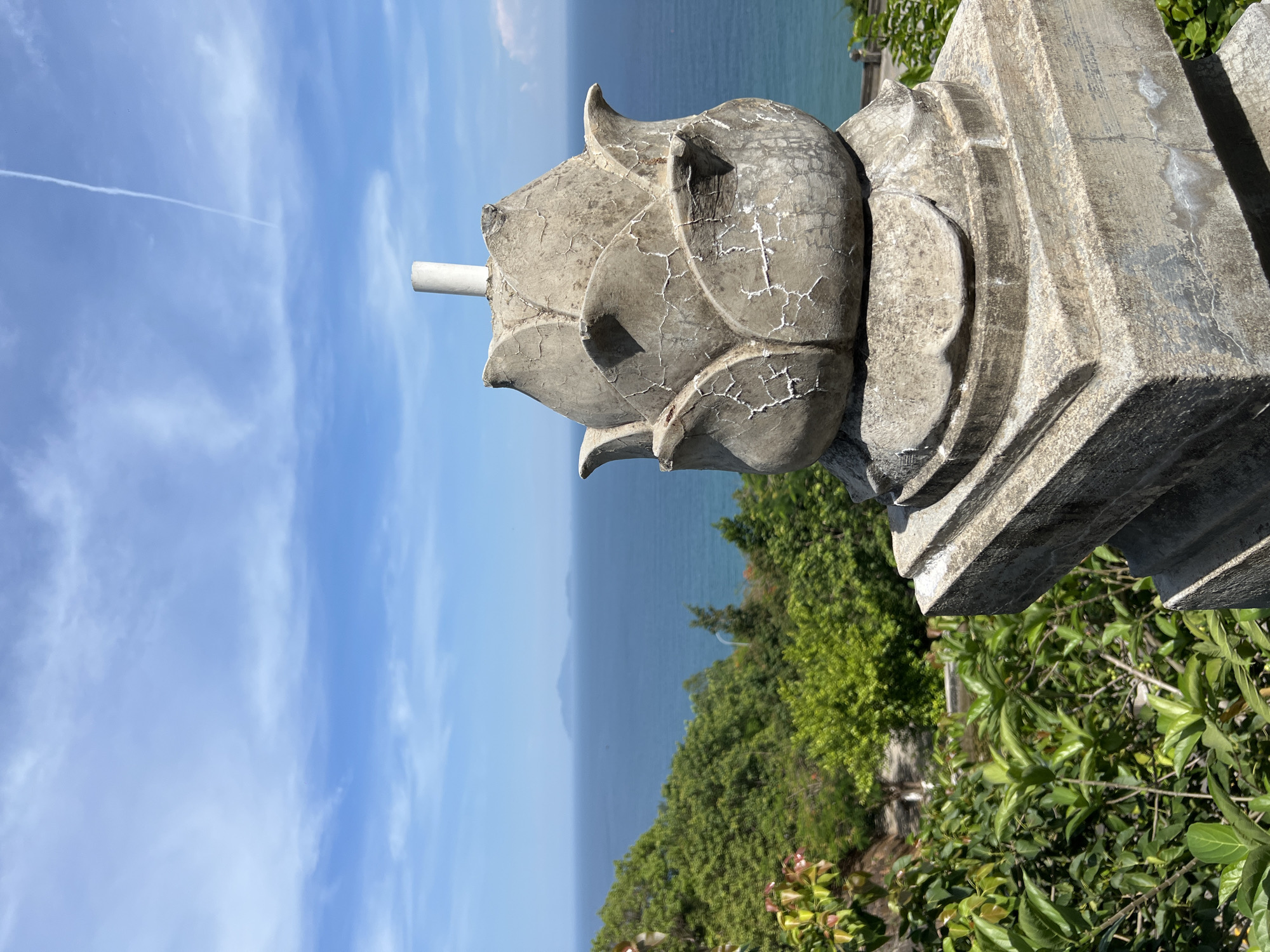 The image captures a close-up of a weathered, lotus-shaped stone carving atop a stone pillar, overlooking a scenic view of the ocean. In the distance, the ocean stretches out towards a hazy coastline with islands. Lush green foliage and trees dominate the foreground and mid-ground, creating a vibrant contrast with the muted tones of the stone sculpture and the blue of the sea and sky. The sky is a clear blue with scattered white clouds and a faint contrail from an aircraft. The stone carving exhibits signs of age and weathering, with visible cracks and a slightly rough texture. The overall setting appears to be outdoors, likely a temple or a viewpoint in a coastal area. Given the context of Đà Nẵng, Vietnam, this scene is likely from a location like the Marble Mountains (Ngũ Hành Sơn), known for its Buddhist temples, caves, and panoramic ocean views, or a similar elevated coastal religious site.