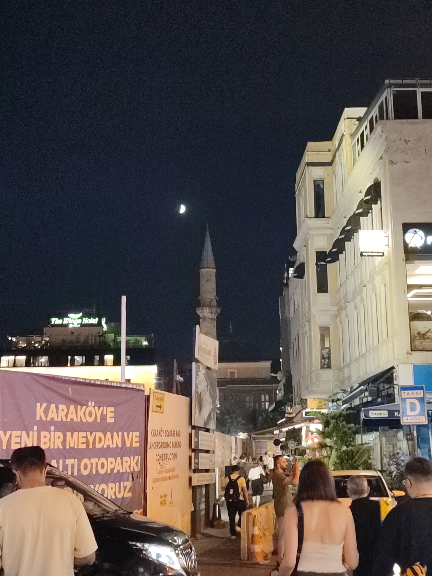 An eye-level, medium shot captures a nighttime street scene in Beyoğlu, Turkey. The sky is dark, with a sliver of a crescent moon visible. A minaret from a mosque rises towards the sky, its thin spire dominating the mid-ground. To the right, a multi-story building with a light-colored facade features numerous windows and architectural details. Signs for "TAKSİ" and "bitaksi" are visible on the building's lower levels.
In the foreground, a large purple banner with yellow Turkish text, including "KARAKÖY'E YENİ BİR MEYDAN VE ALTI OTOPARKI YAPIYORUZ," which translates to "We are building a new square and underground parking lot in Karaköy," is draped across a construction area. Next to it, a yellow sign provides English information about "KARAKÖY SQUARE AND UNDERGROUND PARKING CONSTRUCTION."
Several people are present on the street. On the left, a man wearing a light-colored t-shirt is seen from behind, standing near a dark-colored car. Further down the street, a group of individuals are walking, some carrying backpacks. A yellow taxi is also visible. The overall impression is of a bustling urban environment at night, with ongoing development and pedestrian activity.