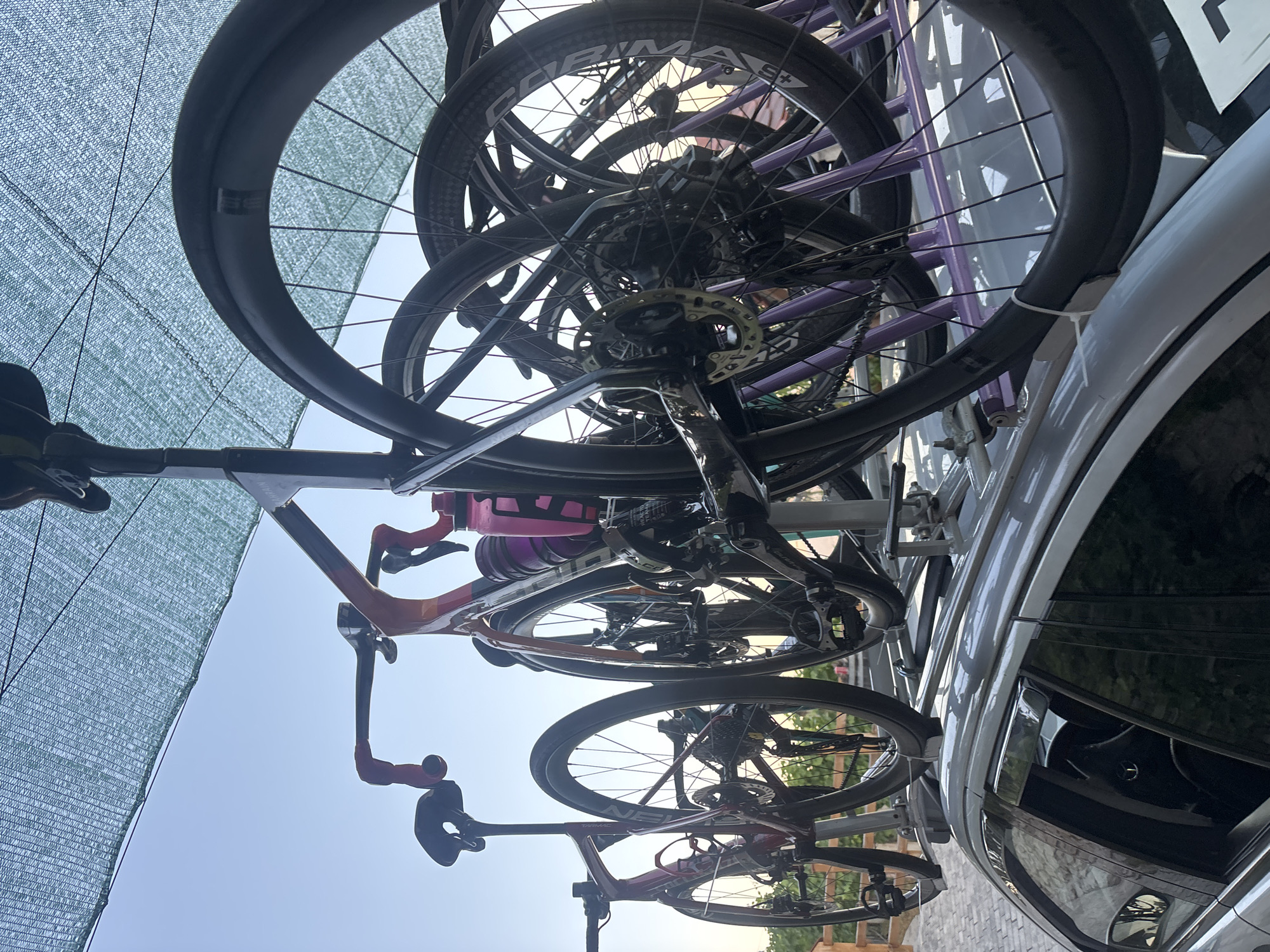 The image depicts a silver car parked on a paved surface, possibly a driveway or parking lot, with a multi-bike rack mounted on its roof. Three bicycles are loaded onto the rack, with their wheels facing outwards. The bicycles appear to be road bikes, featuring thin tires and drop handlebars. One bicycle, in the foreground, is a dark color with red accents on the handlebars and frame. Behind it, another bicycle is visible, predominantly red. A third bicycle, partially obscured, has a frame with orange and pink hues and a pink water bottle attached. The scene is illuminated by natural light, suggesting it is daytime. A green mesh shade covers part of the upper frame, and the sky is visible in the background, indicating an outdoor setting. The overall impression is that of a vehicle prepared for a cycling excursion. The location is specified as Dolenje Grčevje, Slovenia.