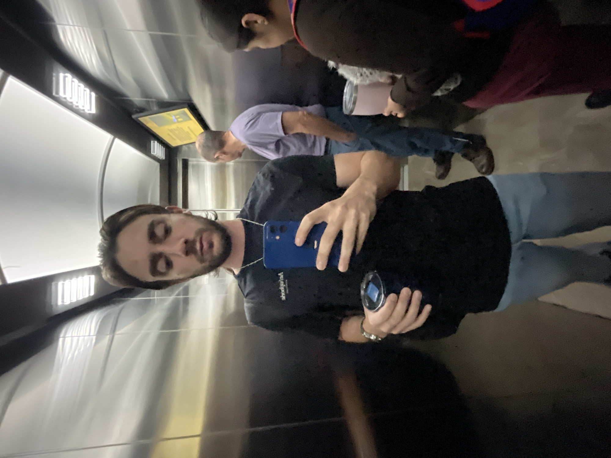 A selfie taken in an elevator, likely in Huixquilucan de Degollado, Mexico, showing a man holding a blue smartphone in his right hand and a tumbler in his left. He is wearing a black t-shirt and blue jeans. Behind him is another man in a purple t-shirt and blue jeans, with brown shoes. To the right of the man in purple is someone with a maroon garment, a black sweatshirt, and a drink. The elevator has stainless steel walls and a control panel to the left of the man in purple. The lighting is bright.