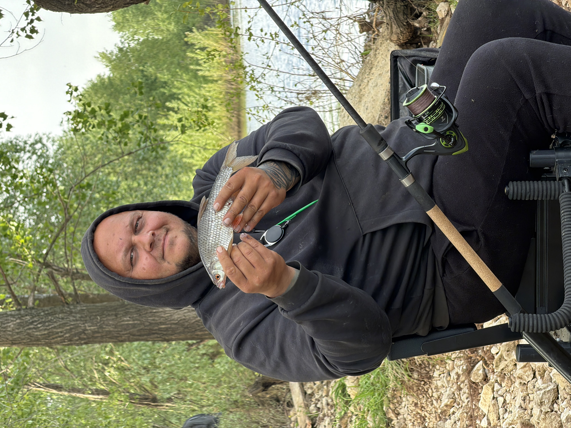 A man is shown seated, holding a fish, likely caught while fishing. He is wearing a dark grey hooded sweatshirt. His right hand, which holds the fish's tail, has a tattoo. The fish is silver with a red fin. A fishing rod is resting on his lap, with a green and black reel attached. He is seated in a fishing chair, and the ground beneath is rocky. In the background, there is a body of water surrounded by trees. The location is most likely in or near Noviy, Russia.