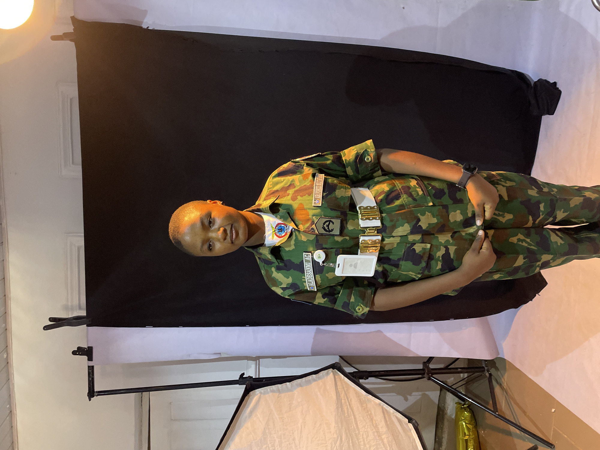 A medium shot of a person in a military uniform with a camouflage pattern, presumably taken in a studio setting. The person is standing in front of a backdrop, which consists of a black fabric draped over a white one. The uniform has badges and patches, including an ID badge. The person is wearing a watch on their left wrist. Studio lighting equipment, including a softbox, is visible in the foreground. The environment appears to be a simple studio setup and there is no indication of the city of Jos, Nigeria, in the image.