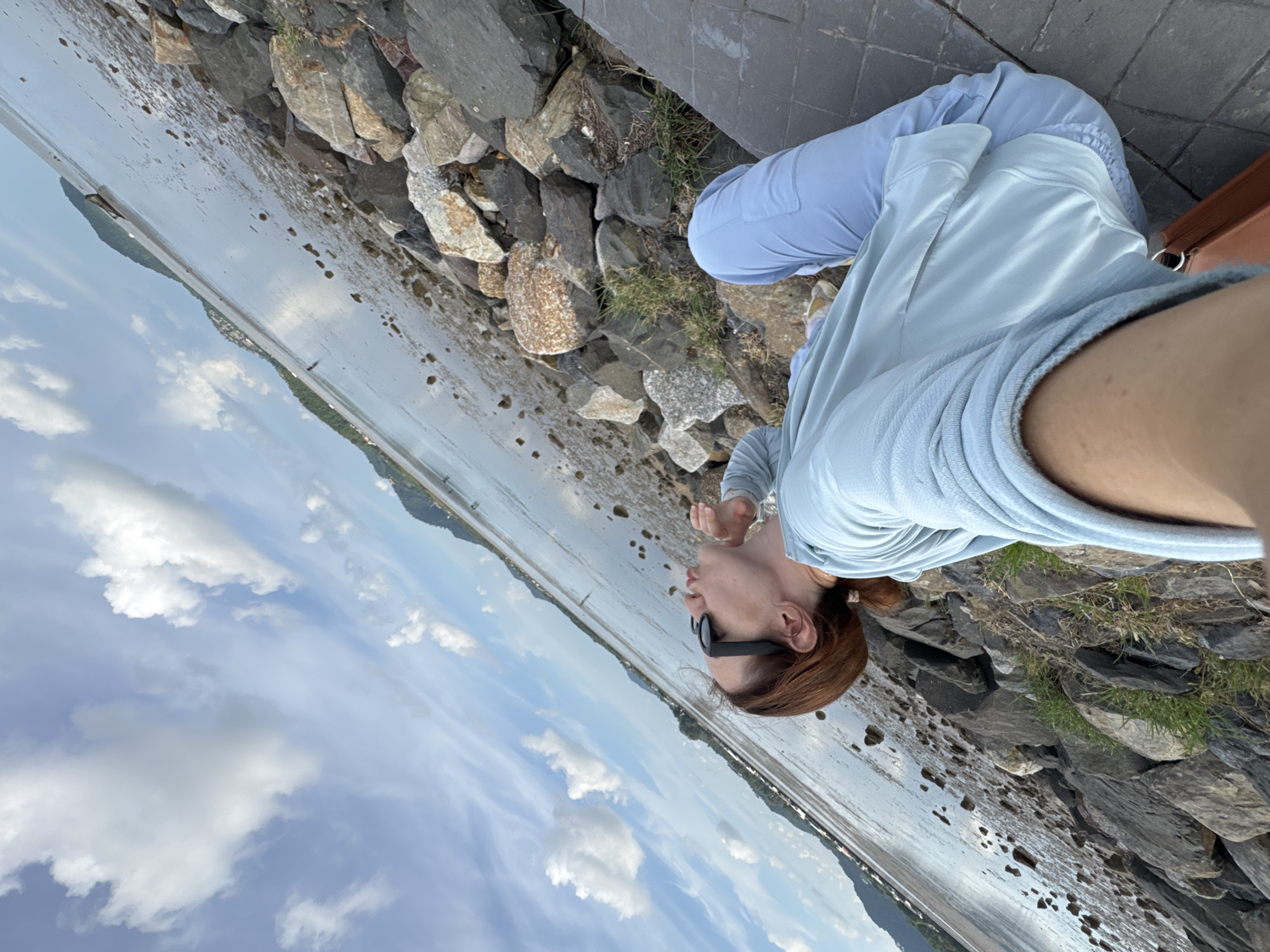 A woman with auburn hair pulled back in a ponytail and wearing sunglasses is posing for a selfie while sitting on a rocky shoreline. She is wearing a light blue hoodie and light blue sweatpants. Her left arm is extended, holding the camera. She is puckering her lips, as if blowing a kiss. The background shows a calm body of water, likely the sea, with islands in the distance. The sky is partly cloudy with a mix of blue and white. The rocky area she is sitting on is covered with various-sized stones and patches of grass. The image appears to have been taken outdoors on a clear day. The location is described as the city of Phuket City Municipality, Thailand.