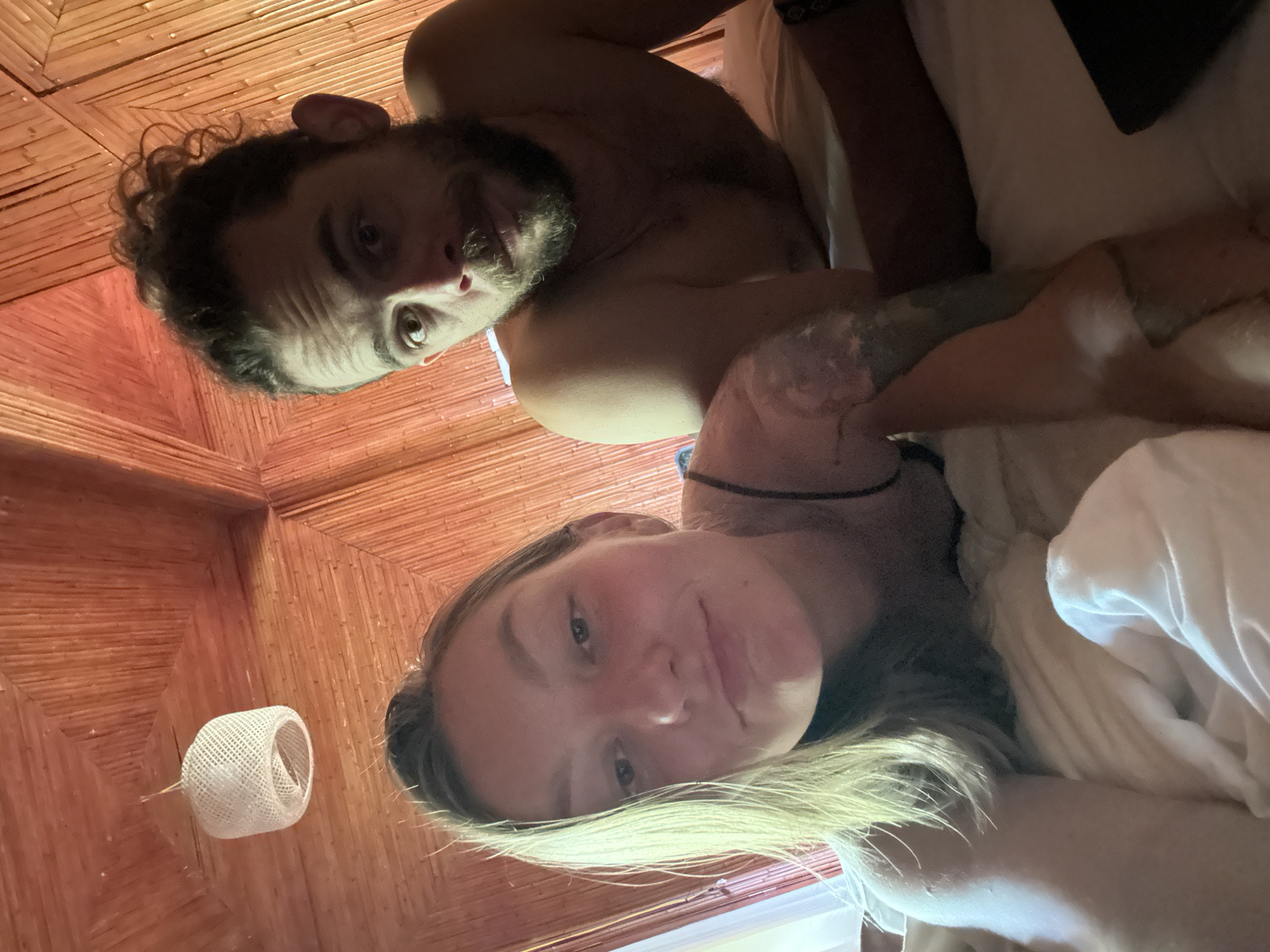 This vertical selfie captures two individuals, a man and a woman, lying down and looking up towards the camera. The setting appears to be an indoor space, likely a bedroom, characterized by a distinctive ceiling or wall constructed from light brown or reddish-brown woven bamboo or reed panels, arranged in a geometric, radiating pattern.
The man, positioned closer to the camera, has dark, curly hair and a beard. He is shirtless, revealing body hair and tattoos on his left arm. He gazes directly at the camera with wide eyes and a neutral expression.
Behind and slightly to the side of the man, the woman is partially visible. She has long, blonde or light brown hair that cascades around her face and shoulder. She has a fair complexion and a soft, slight smile as she looks towards the camera. Both individuals are partially covered by white bedding or sheets, reinforcing the impression that they are in bed.
A small, white, cylindrical decorative object, possibly a woven light fixture or lantern, hangs from the bamboo ceiling in the upper left background. The overall lighting is dim and warm, suggesting an intimate indoor atmosphere. No text is visible in the image. The architectural style, particularly the bamboo paneling, is consistent with traditional or rustic designs found in Southeast Asia, aligning with the given location context of Thông Nguyên Commune, Vietnam.