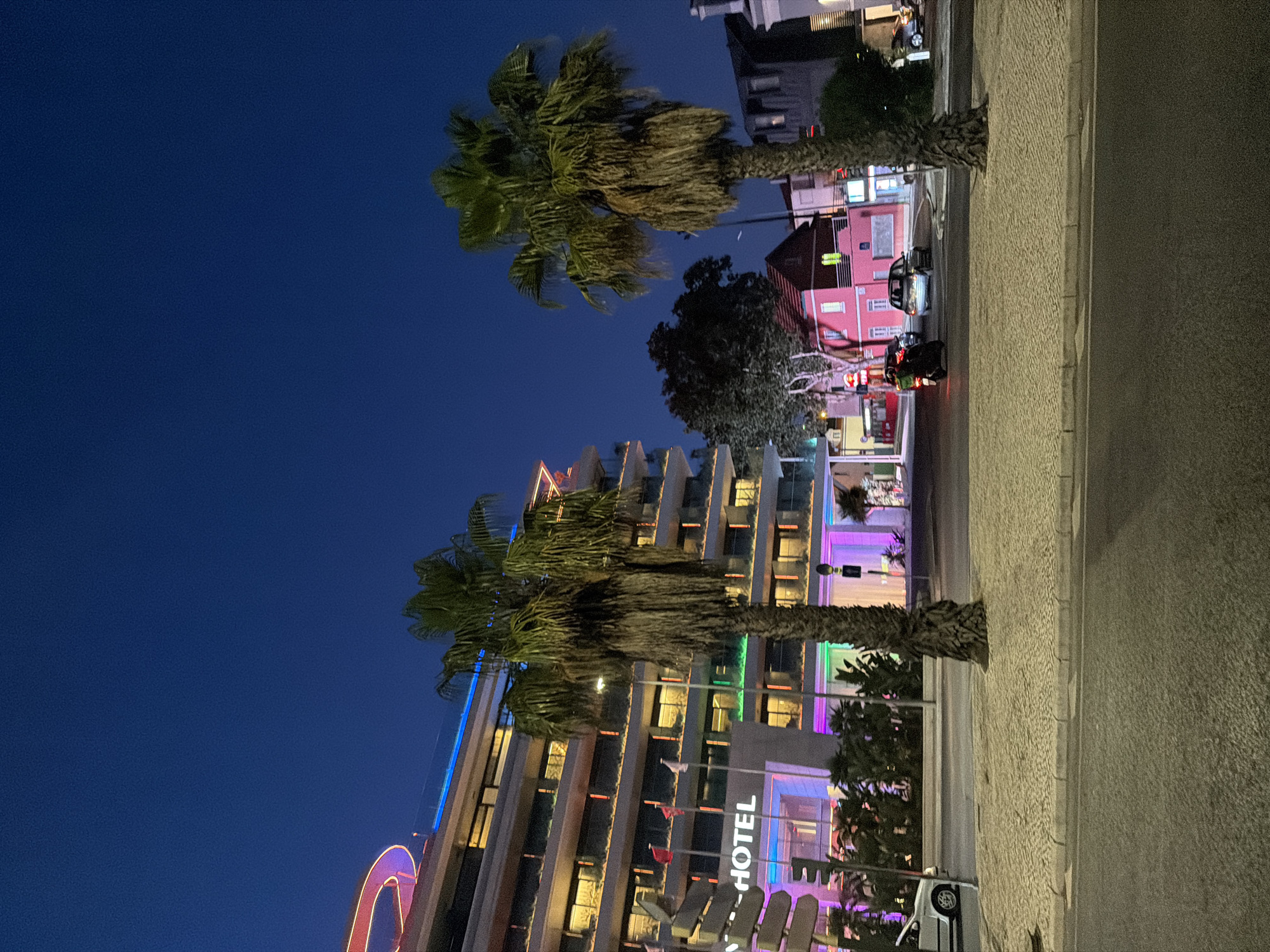 This media file depicts a nighttime scene in Cascais, Portugal. The image features a modern hotel building with illuminated windows and colorful exterior lighting, including blue and purple hues. Palm trees are prominent in the foreground, their fronds swaying, suggesting a breeze. A sidewalk with a textured surface runs alongside a road where a few vehicles, including a motorcycle and a car, are visible. In the background, other buildings, including a pink one, are also lit up. The sky is a deep blue, indicating it is nighttime. The overall atmosphere suggests a lively, urban setting, possibly a resort town or a vibrant city street. There is no clear indication of people engaging in specific activities, but the presence of vehicles and illuminated buildings points to ongoing activity. Visible text includes "_HOTEL" on the hotel building. The time of day is clearly night, and the weather appears clear.