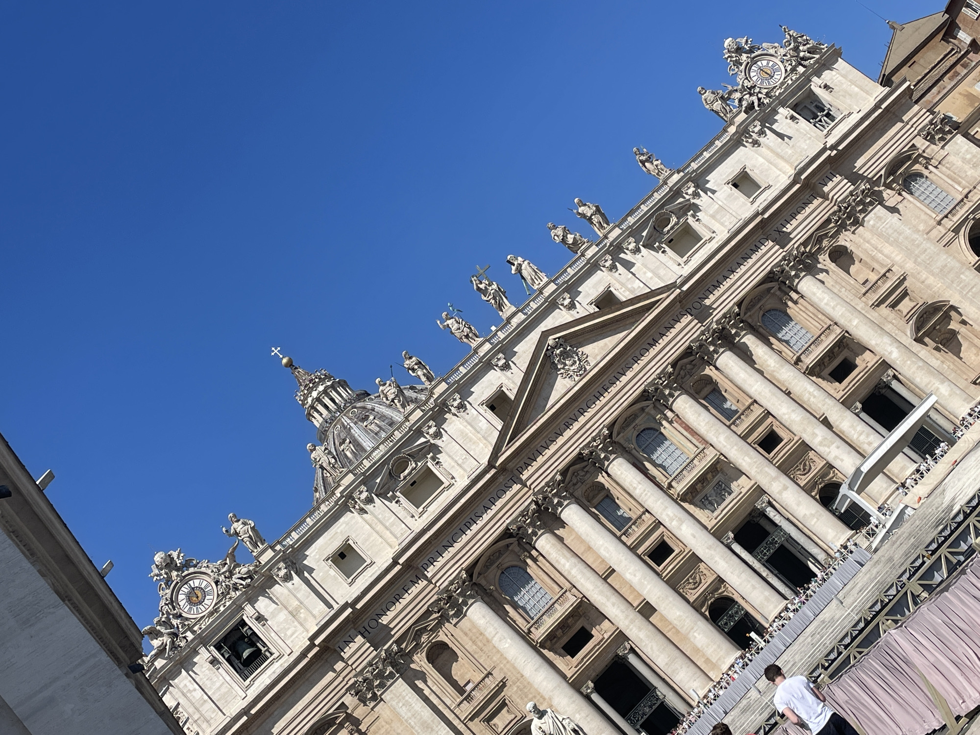 The image shows a low-angle view of a section of St. Peter's Basilica in Città del Vaticano, Italy. The building's facade is richly decorated with sculptures, columns, and windows. A dome is visible in the background. Statues line the top of the facade. A clock is visible near the building's top edge. The sky is a clear blue. A temporary structure with a fabric covering is visible at the bottom of the frame, with people standing on it. One person is visible wearing a white t-shirt.