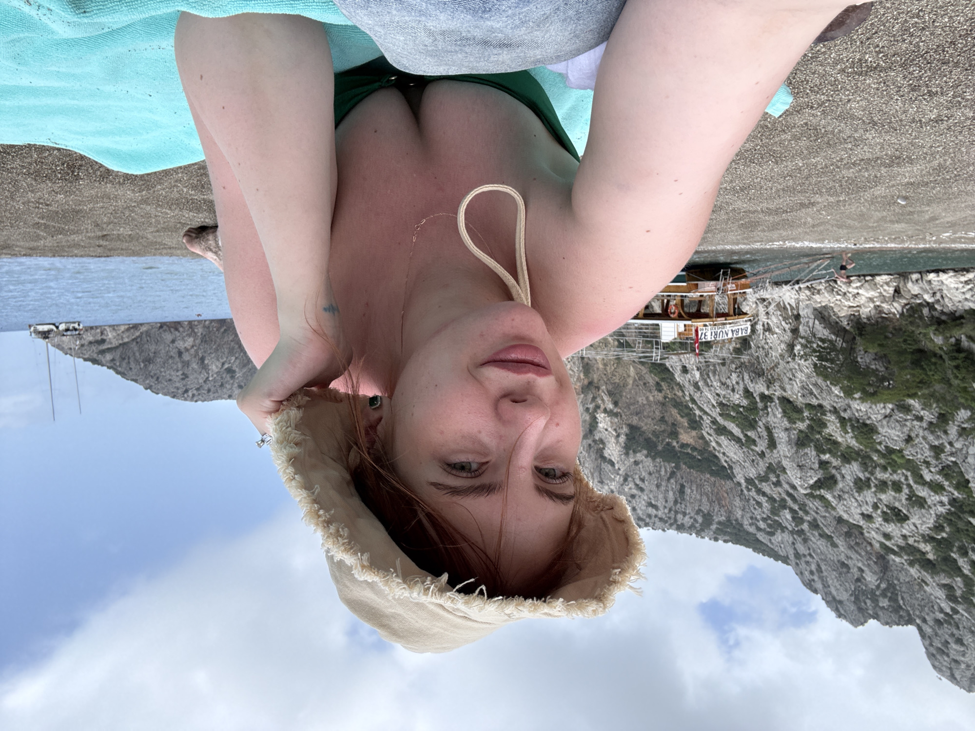 The image depicts a woman with fair skin, wearing a green swimsuit and a beige sun hat, who is likely taking a selfie. The background shows a coastal scene with a rocky mountain range, a boat labeled "BABA NUR 37" in Turkish, and the sea. The location is likely in Kumluca, Turkey, given the boat’s Turkish inscription. The woman is situated on a beach or pier, indicated by the concrete surface and sand. There is a blue towel under her. The weather appears sunny with some clouds.