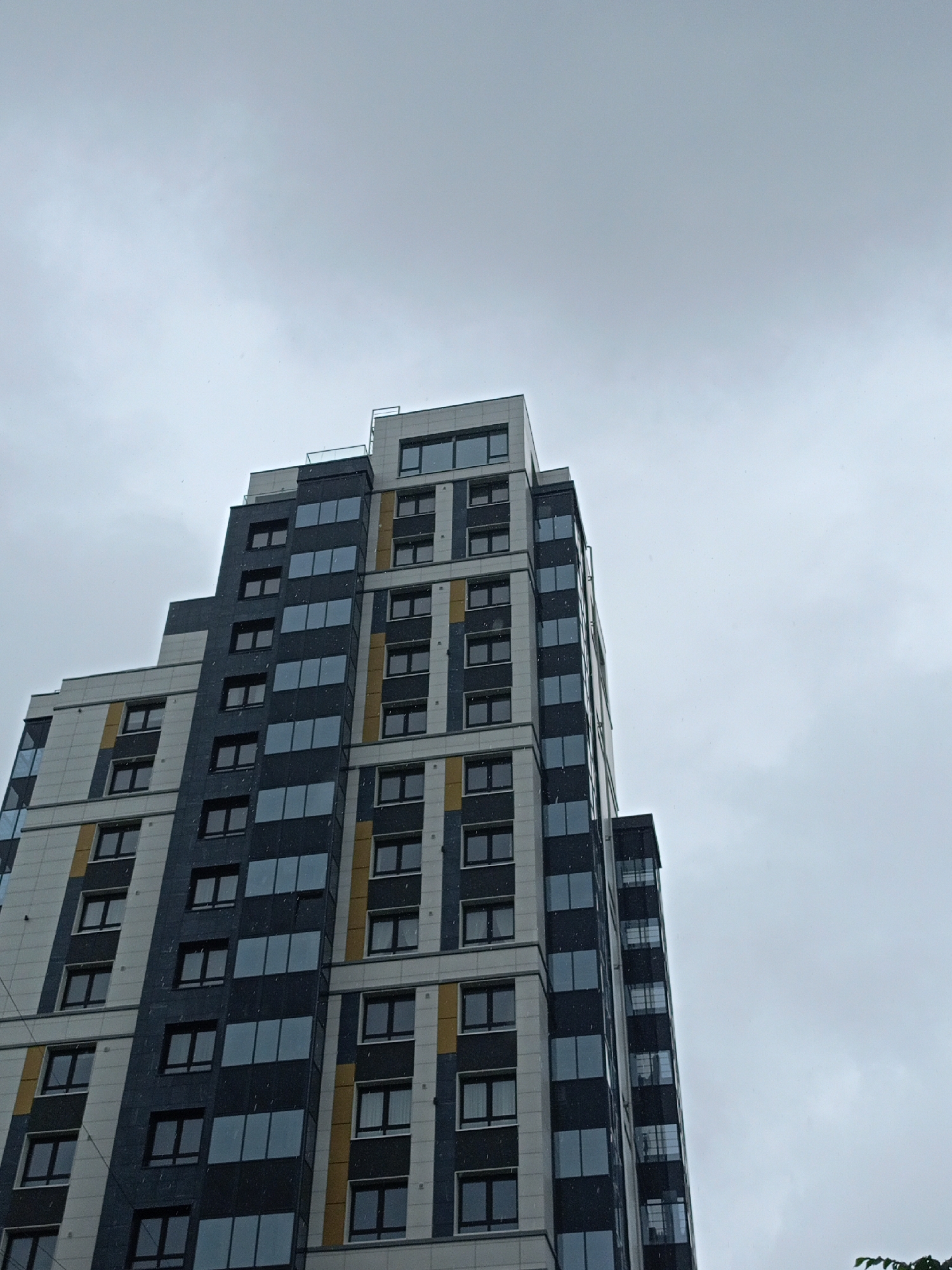 The image shows the exterior of a modern, multi-story apartment building. The building has a distinctive design with alternating sections of light-colored cladding and dark grey panels with numerous windows. There are also accents of yellow in some sections, breaking up the monochrome. The windows are mostly rectangular and appear to be tinted or reflective. The building's architecture is geometric, with some staggered levels and protruding sections.
The scene is dominated by the building which takes up the majority of the frame, angled upwards towards a cloudy, overcast sky. The sky is a uniform grey, suggesting a possibly dull or rainy day. There are some small, white specks visible in the air, which could be snow or rain. No people are visible in the image, and the focus is solely on the architectural structure. The setting appears to be urban, given the nature of the building. No text is visible in the image. The overall mood is somewhat somber due to the weather and lighting conditions, emphasizing the building's imposing structure against the muted sky.
