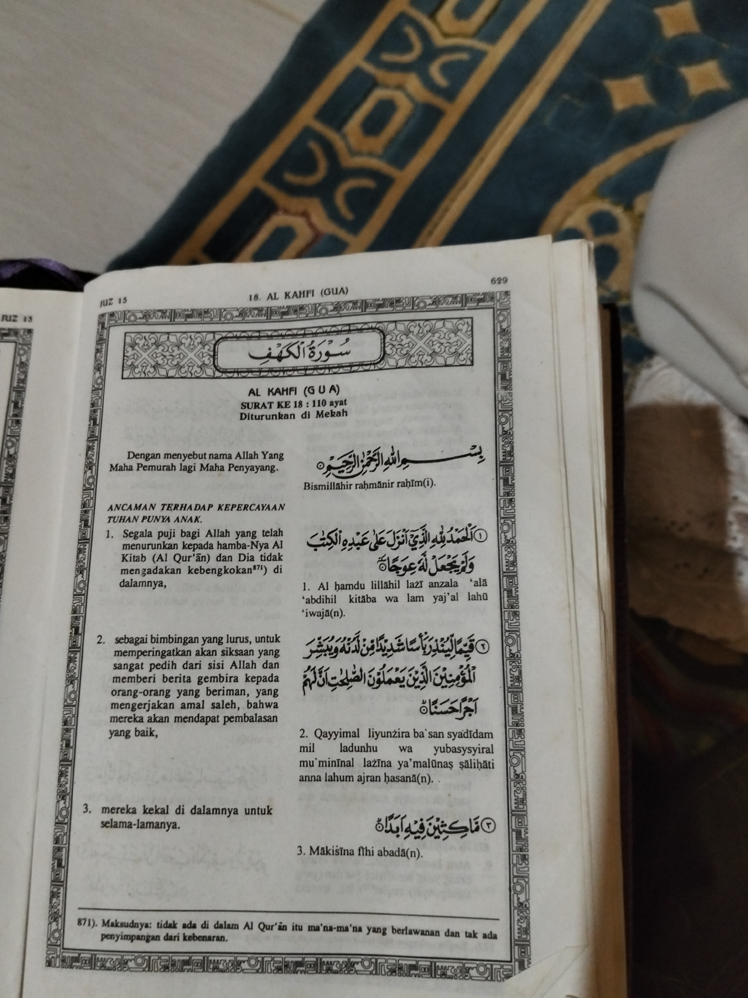 The image shows an open book, likely a Quran, with Arabic text and its Indonesian translation. The book is placed on a patterned rug. The page displays Surah Al Kahfi (Gua), Verse 18, with 110 verses, which was revealed in Mecca. The text includes the Basmalah and verses discussing divine attributes and warnings. The context of the image's location in the city of Payagaboh, Indonesia is not depicted or discernible within the provided visual information. The image focuses solely on the book and its contents, with a portion of a rug visible in the background.