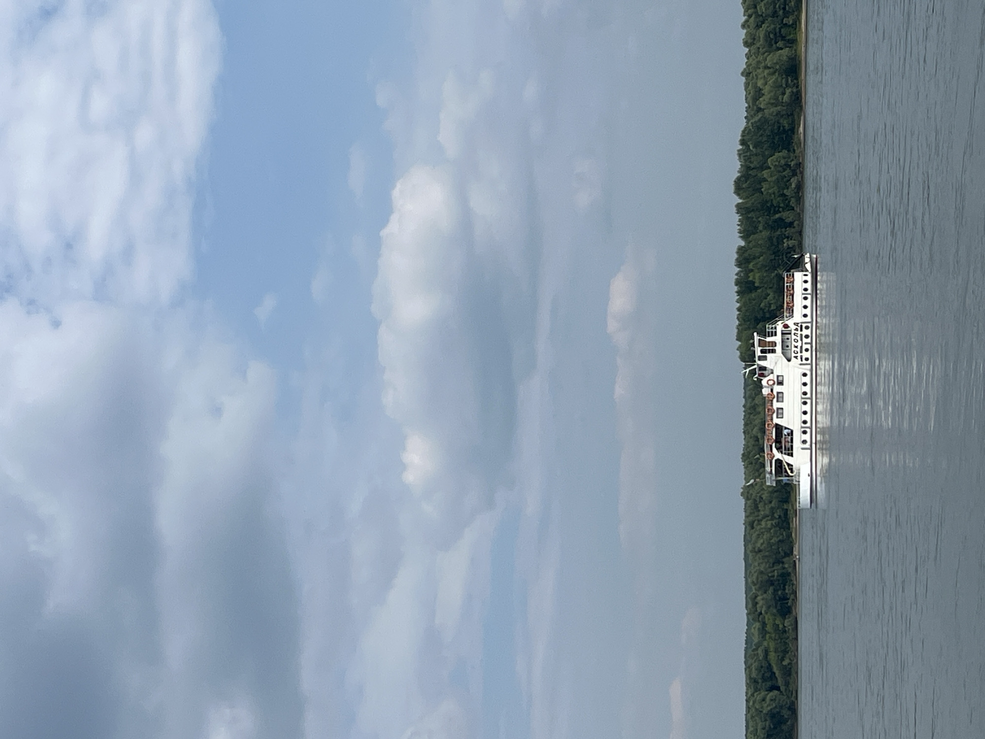 A white passenger boat with red trim is traveling on a wide, calm river. The boat has multiple windows and decks, and life preservers are visible on its railings. The river is bordered by a dense, green forest on the horizon. The sky is partly cloudy with patches of blue. The scene appears to be located in Mochishche, Russia, where the river likely flows. There are no people visible in the image.