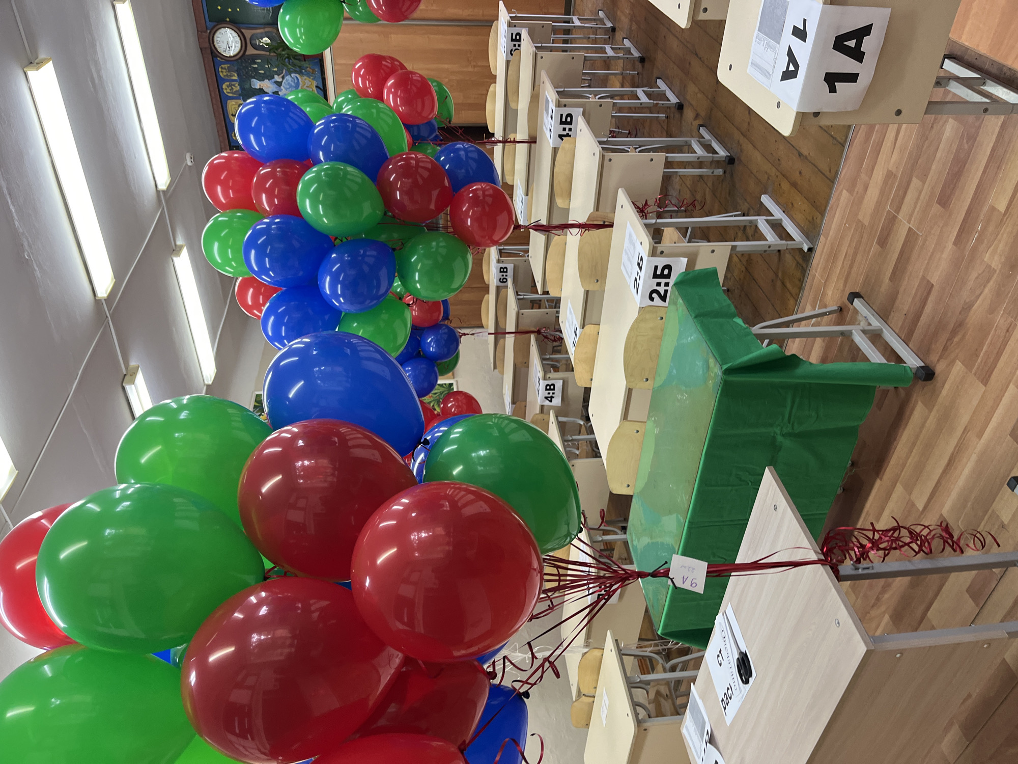 The image depicts an interior space, likely a classroom, decorated with red, green, and blue balloons. School desks are arranged in rows. Each desk has a tag with a number and letter combination. A green cloth covers one of the desks. The photograph was taken in Kaspiysk, Russia.