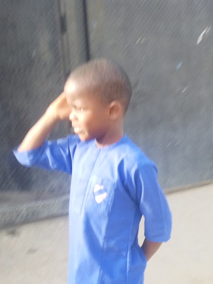 A young child, with short, dark hair, is depicted in Garun Gawa, Nigeria. The child wears a blue, three-quarter sleeve tunic-style garment, featuring a light-colored, shield-shaped emblem with blue horizontal stripes on the left chest pocket area. The child's right hand is raised, with fingers positioned near the right temple/forehead. The child's head is turned to the right profile, and the mouth is slightly ajar. The background consists of a dark, textured wall surface with a vertical dark line visible on the far left.