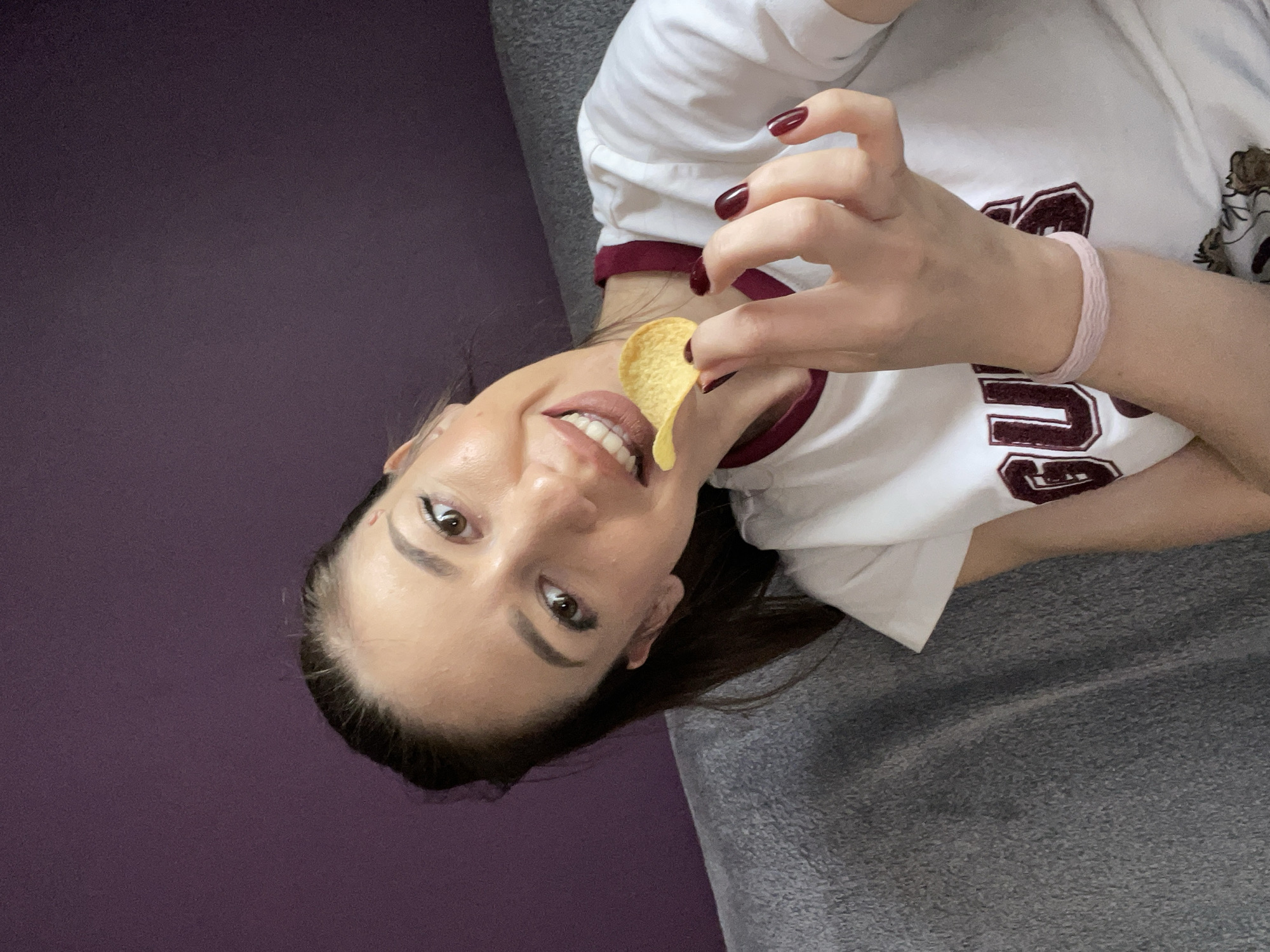 A female individual is positioned at an angle in the image, holding a potato chip up to her mouth. She has fair skin, brown eyes, and dark hair pulled back. Her fingernails are painted a dark shade of red. She is wearing a white t-shirt with a maroon trim and lettering. A light pink bracelet is visible on her left wrist. She appears to be lying on a grey fabric surface with a purple wall in the background. The location of the image within the city of Sofia, Bulgaria cannot be determined based on the available visual information.